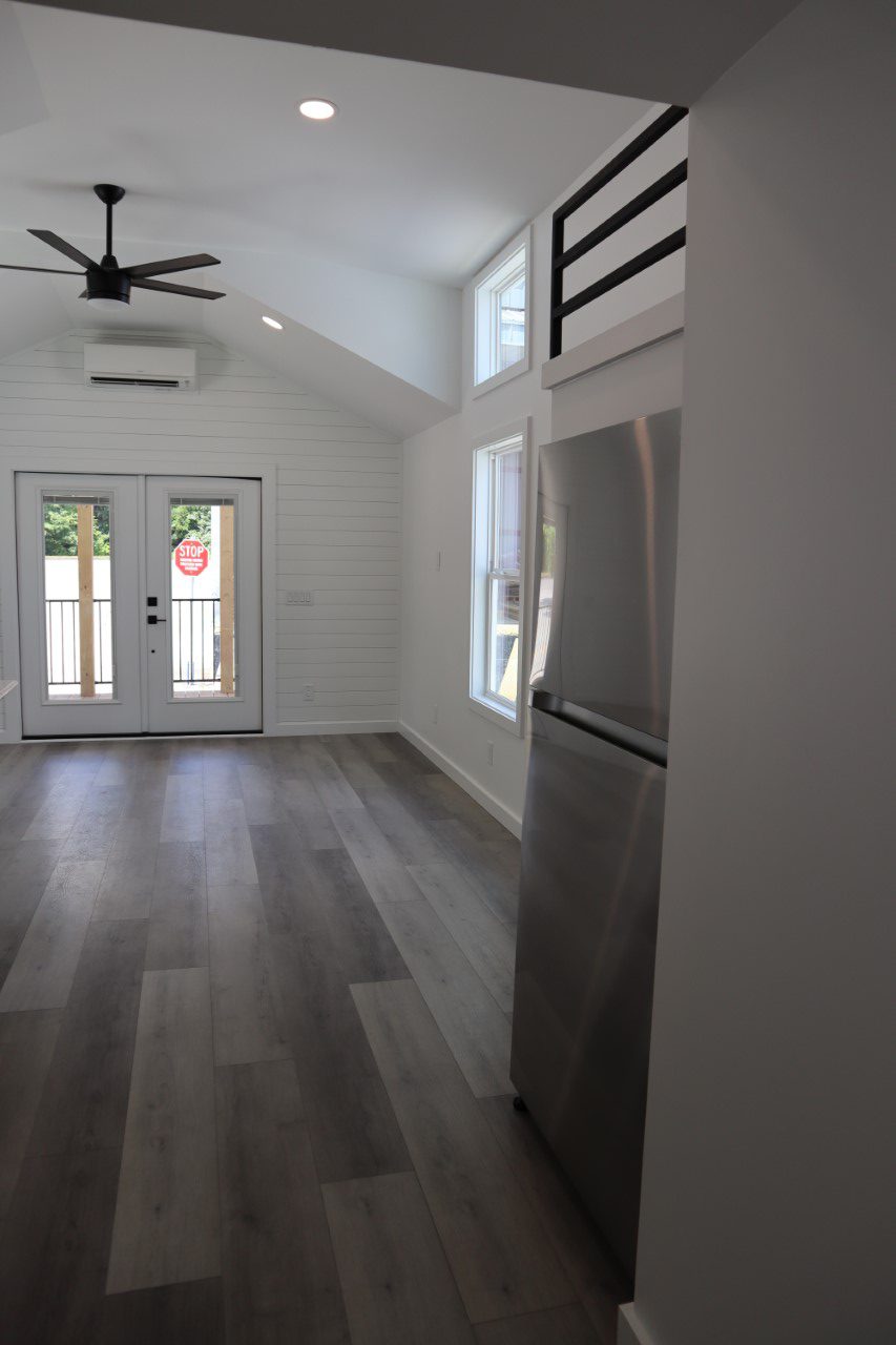 Modern, minimalistic room with gray wood flooring, white walls, ceiling fan, and large windows. A stainless steel refrigerator is to the right.