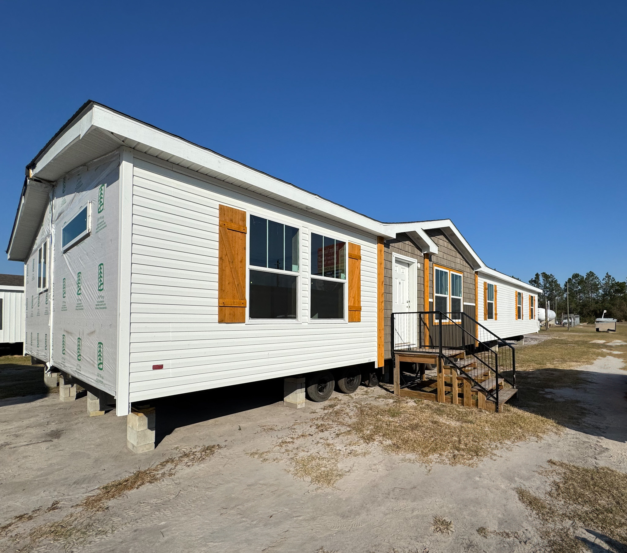 A white manufactured home with wood shutters and a small porch sits elevated on blocks against a clear blue sky, creating a peaceful, sunny rural scene.