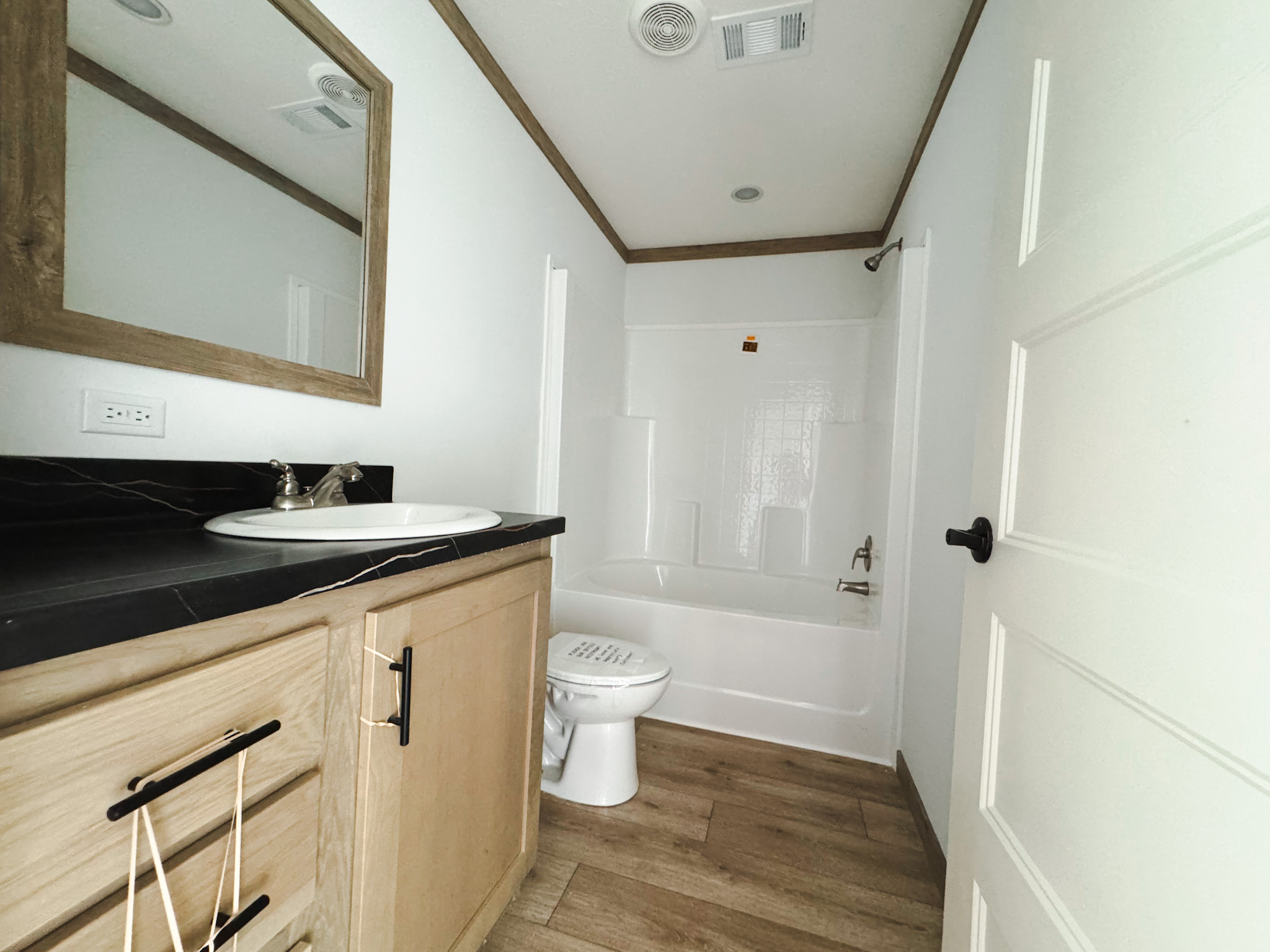 Modern bathroom with light wood cabinets, black countertop, and white sink. A bathtub and toilet are in the background, with wood flooring throughout.
