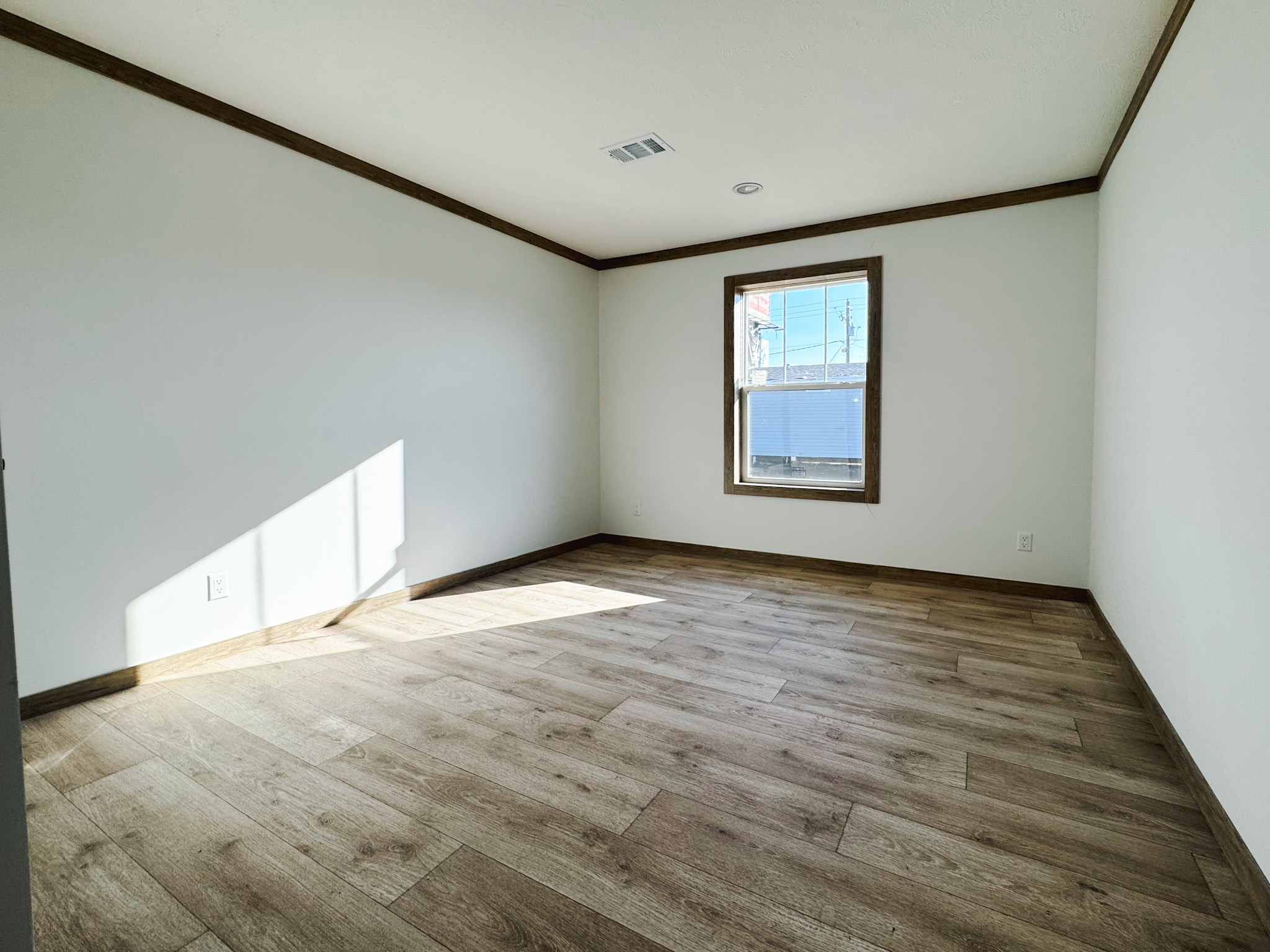 A sunlit, empty room with white walls and brown wood floors. A single window on the right wall frames an outdoor view. Calm, minimalist atmosphere.