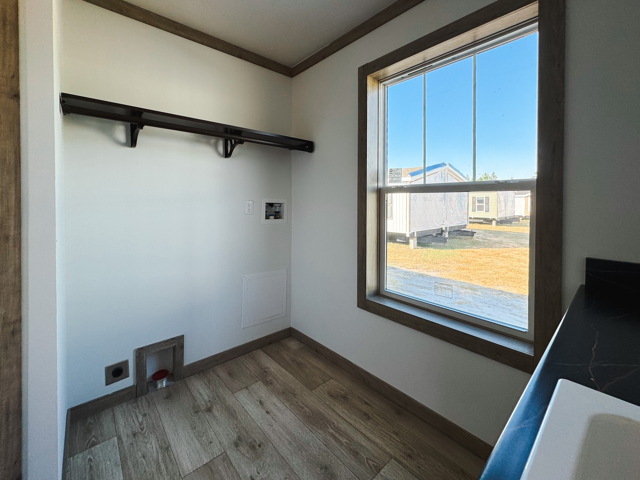 Compact laundry room with wood floor and beige walls, featuring a black shelf. Sunlight streams through a window, offering a view of nearby houses.