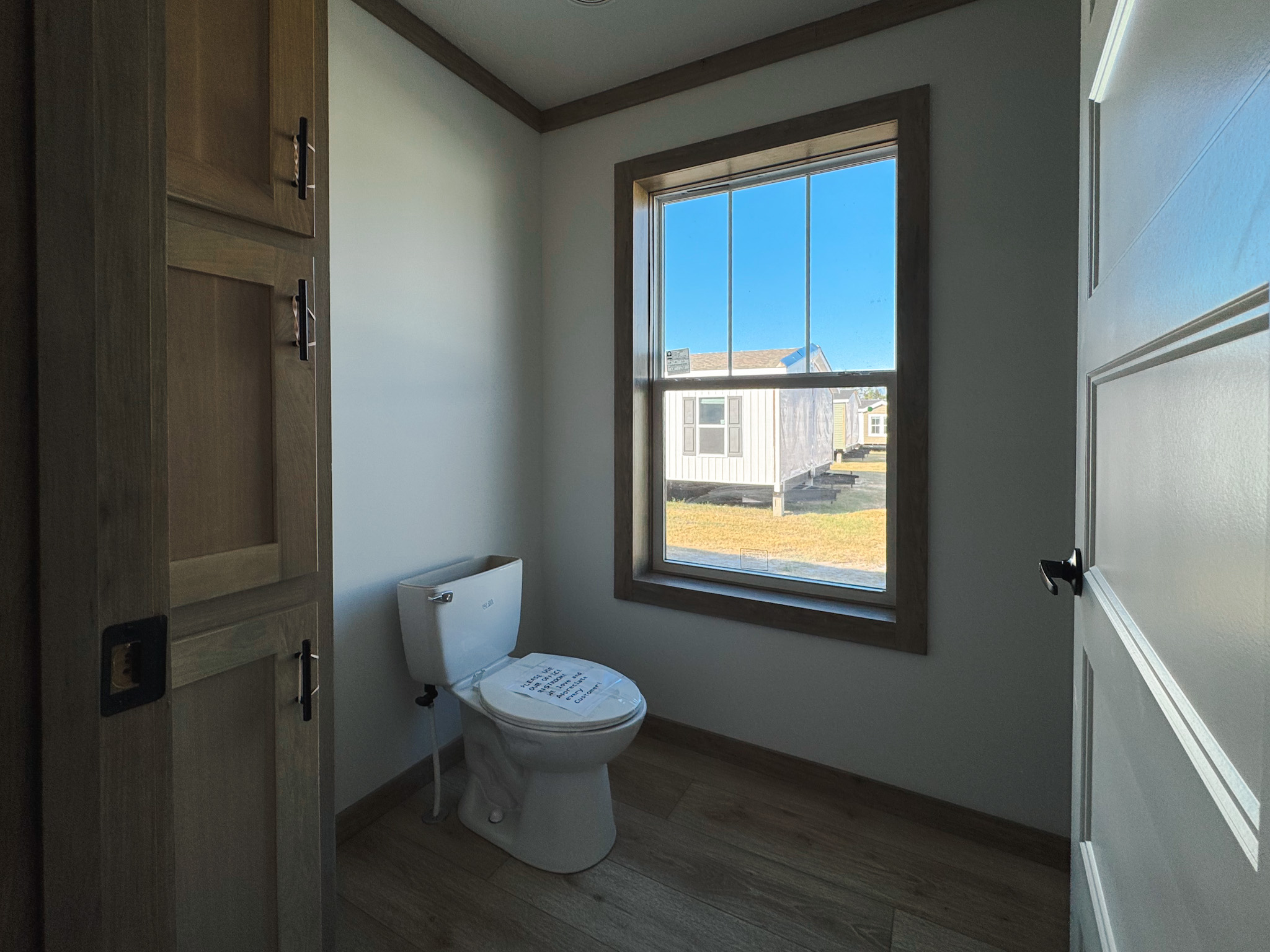 Small, simple bathroom with a wooden cabinet, white walls, and a toilet under a large window showing a sunny outdoor scene with mobile homes.