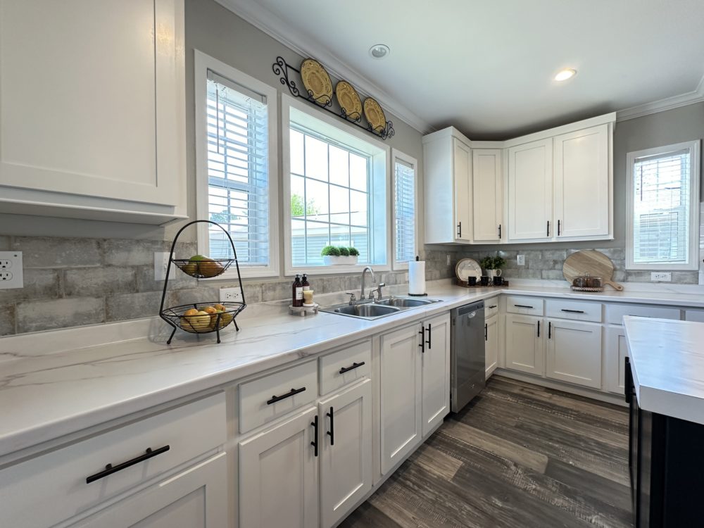 Bright kitchen with white cabinets, marble countertops, and stainless steel sink. Natural light pours through large windows, creating a cozy atmosphere.