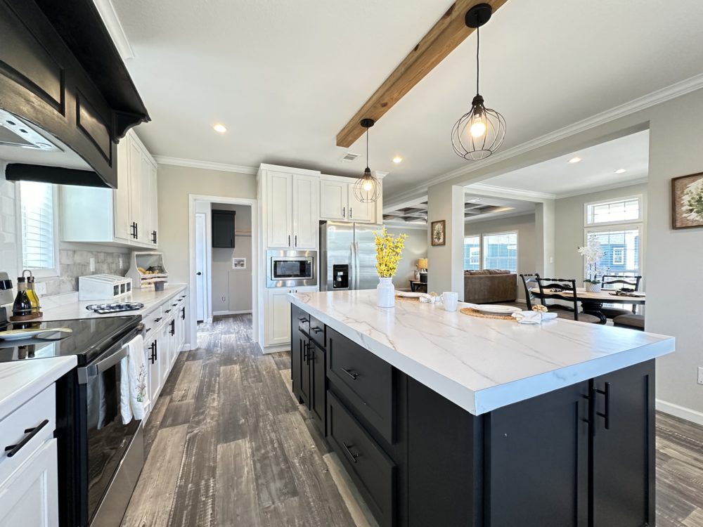 Modern kitchen with a white countertop island and black cabinets, pendant lights, stainless steel appliances, and yellow flowers in a vase, opening to a bright living area.