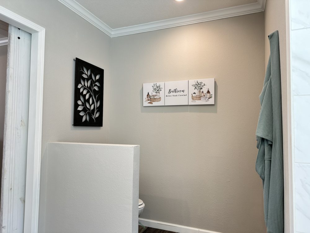 Bathroom corner with a decorative leaf wall art and a calming "Bathroom" sign above. A teal towel hangs on the right. Minimalistic and serene.