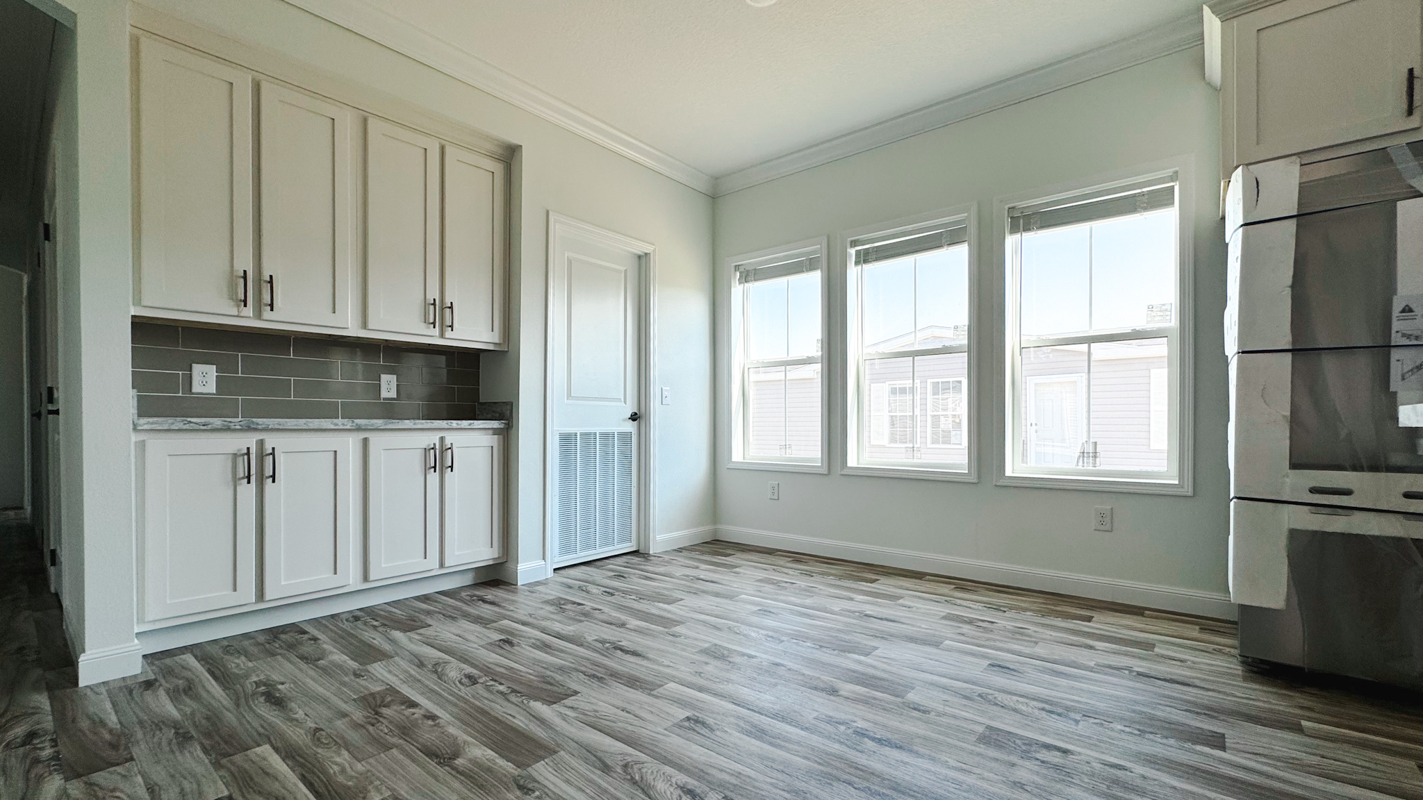 Bright kitchen with light wood flooring, white cabinets, and gray backsplash. Three large windows on the right let in natural light, creating a fresh atmosphere.