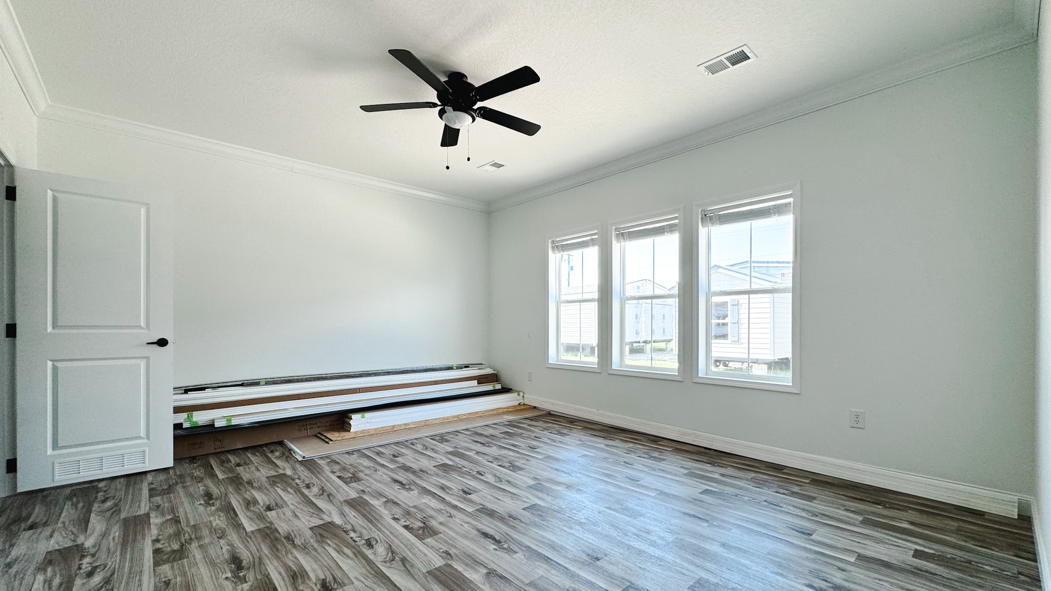 A bright, empty room with light gray walls, three tall windows, and wood-look flooring. A ceiling fan is centered, and wood planks are stacked on one side.