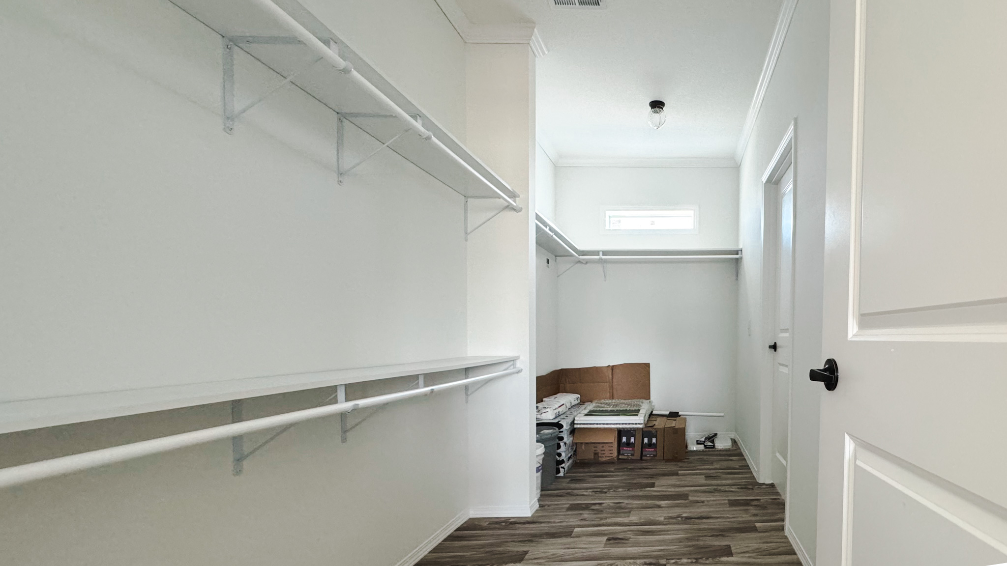 Spacious walk-in closet with white walls, ceiling-high shelves, and hanging rods. Wooden floor adds warmth. Boxes are stacked at the far end.