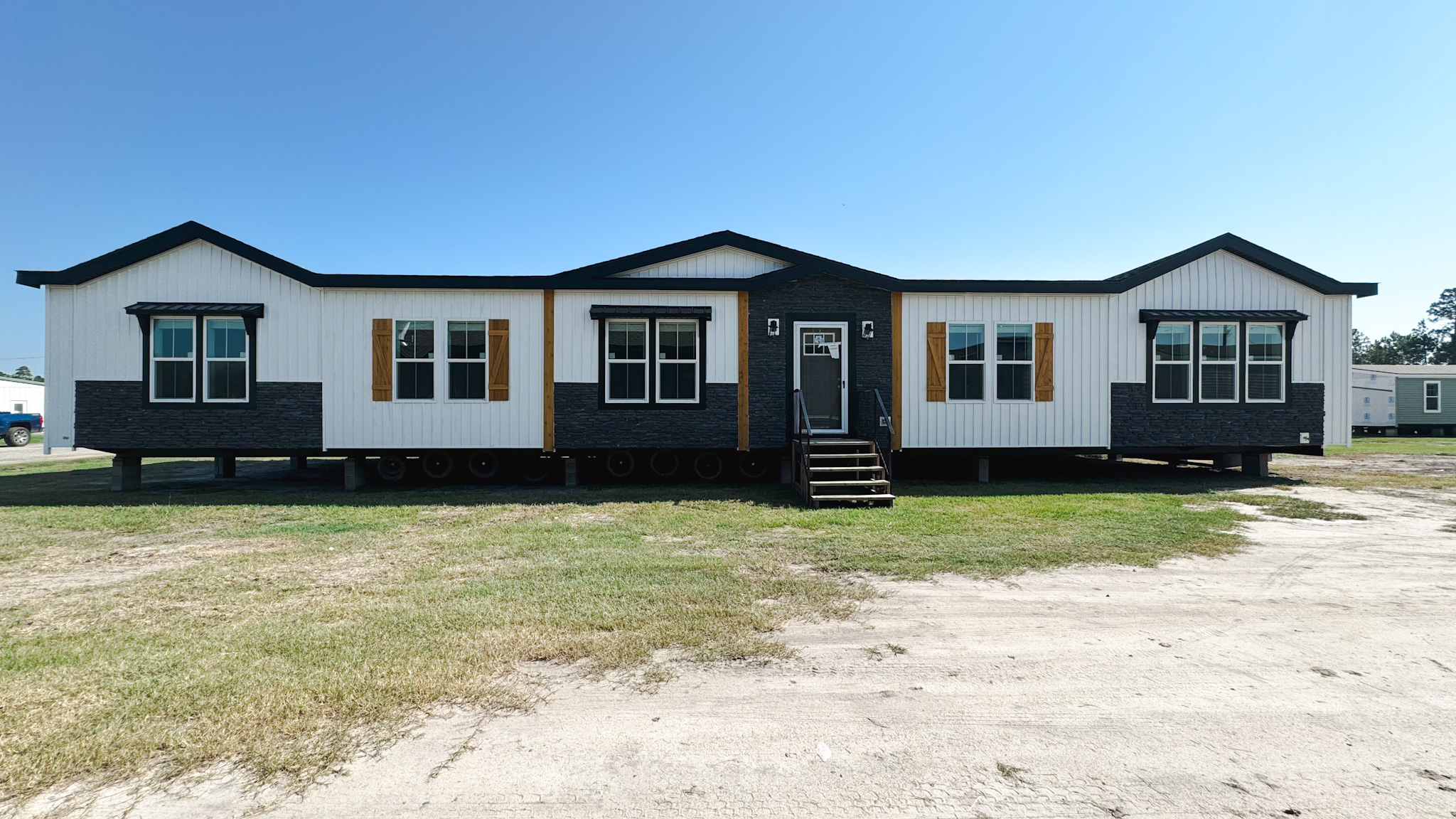 A modern, double-wide modular home with a black and white exterior, wooden accents, and multiple large windows sits in a grassy area under a clear blue sky.
