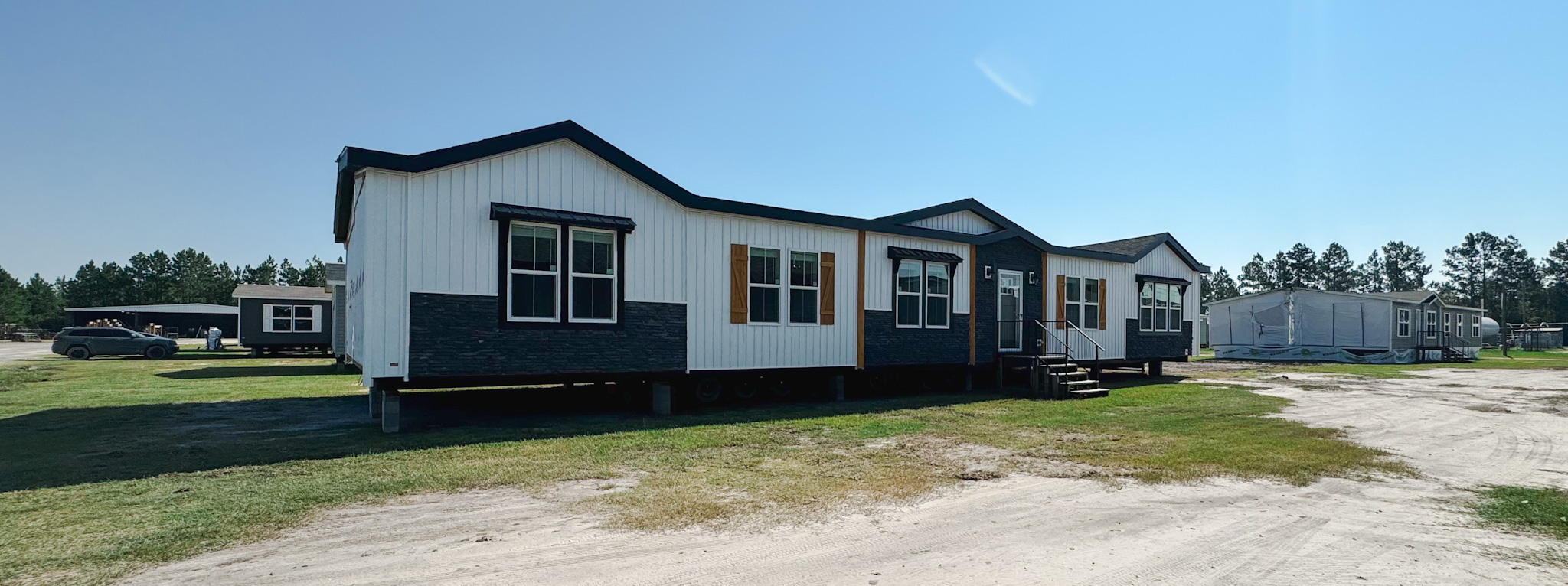 A long, white and black modular home with a pitched roof sits on grassy terrain under a clear blue sky. Other modular homes are visible in the background.