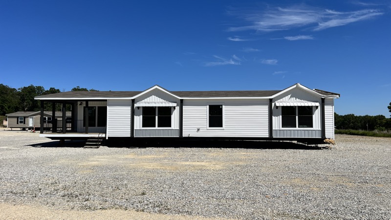 A single-story, white manufactured home with a gray roof sits on a gravel lot. The home features large windows and a small porch, under a clear blue sky.