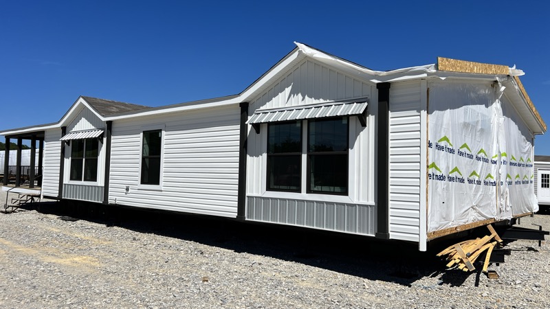 A partially constructed house with white siding, two large front windows, and a covered section with tarpaulin. Set on a gravel lot under a clear blue sky.