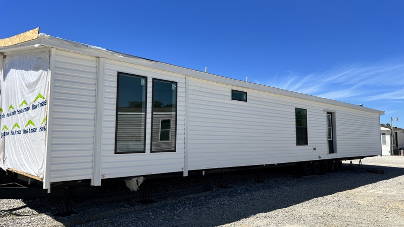 A long, white mobile home is parked on a gravel lot under a clear blue sky. It features large windows and is wrapped in protective material on one end.