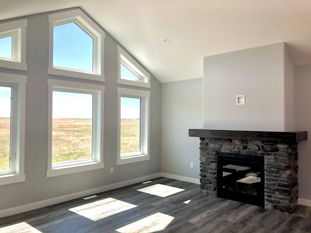 Modern living room with high ceilings, featuring large windows allowing natural light, a stone fireplace with a dark mantel, and wood floors. Calm ambiance.