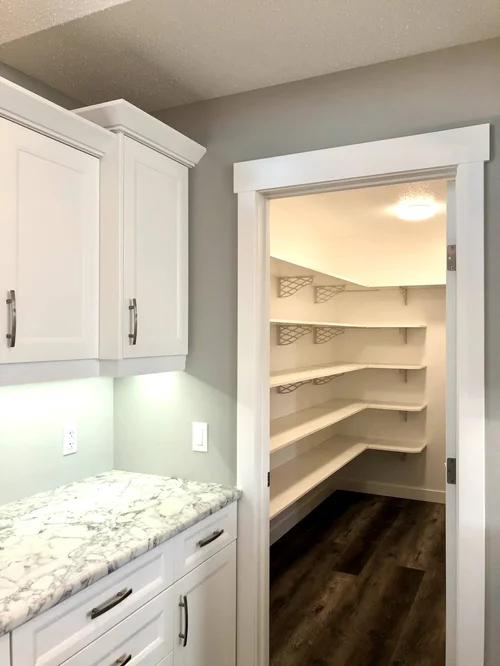 Bright pantry with white shelves and gray walls, adjacent to a kitchen with white cabinets and a marble countertop. The space feels modern and organized.