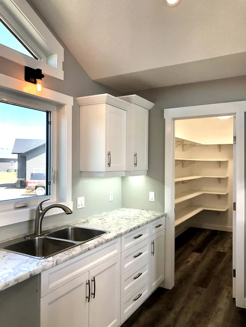 Bright kitchen with white cabinets and marble countertops. A window above the sink allows natural light. Open door reveals a pantry with wooden shelves.