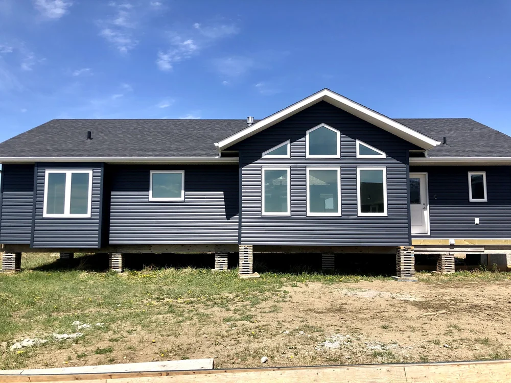 A blue modular house on stilts under a clear sky. Features include large windows and a gabled roof. The ground is bare, suggesting recent construction.