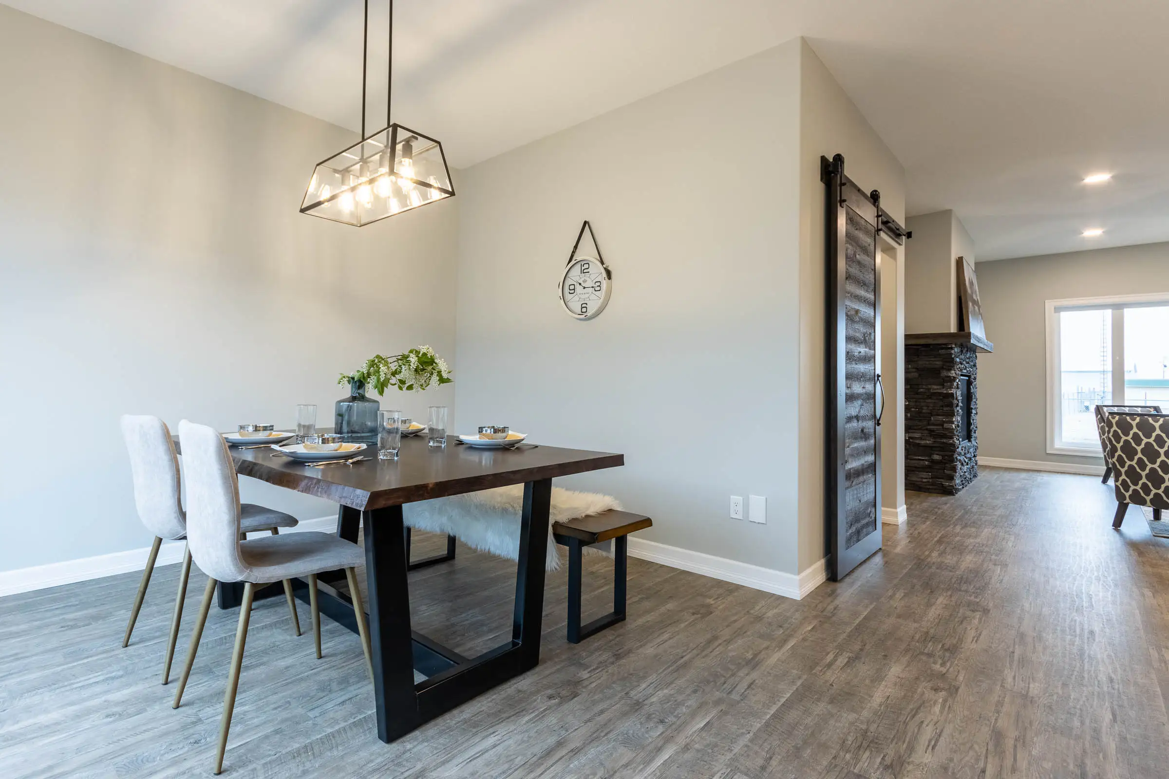 Modern dining area with a wooden table, four grey chairs, and a bench on a light wood floor. A geometric pendant light and wall clock add elegance.