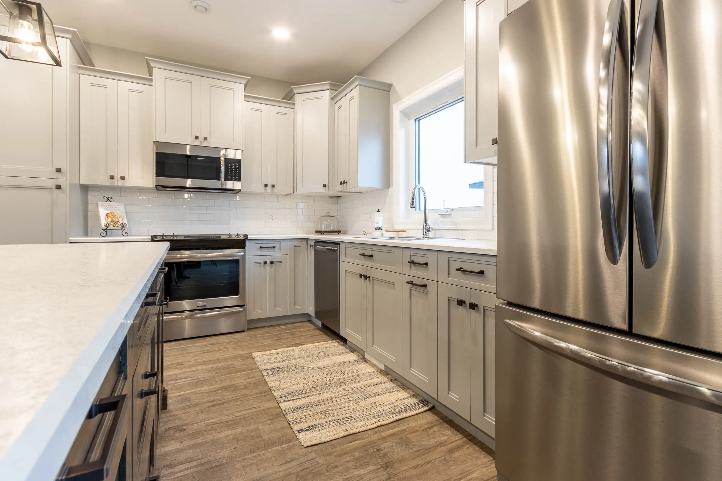 Modern kitchen with light gray cabinets, stainless steel appliances, and a wooden floor. A woven rug adds warmth. Bright, clean, and inviting.