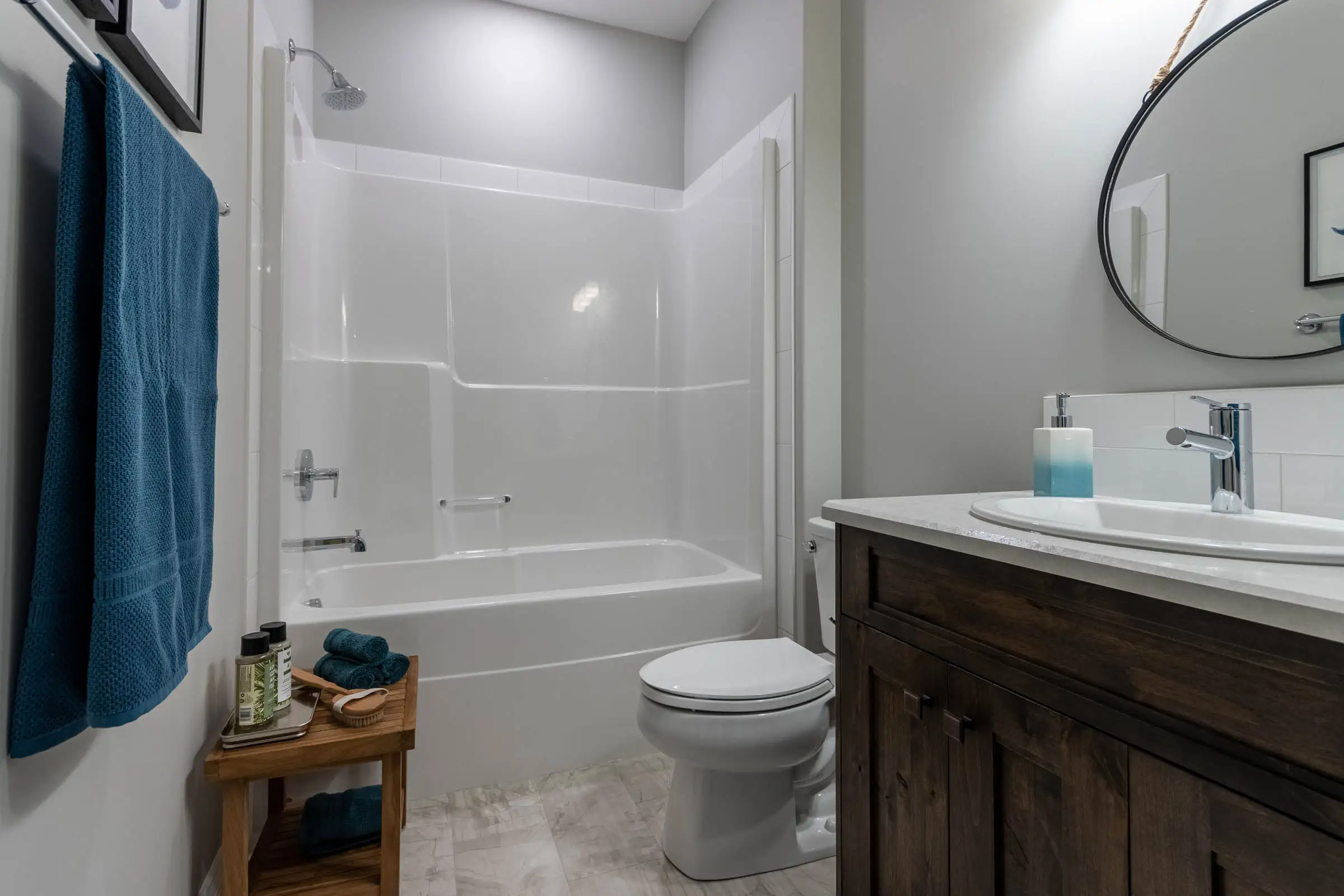A modern bathroom with a white bathtub and shower, wooden vanity, round mirror, and blue accents like a towel and bottles, creating a clean, cozy vibe.