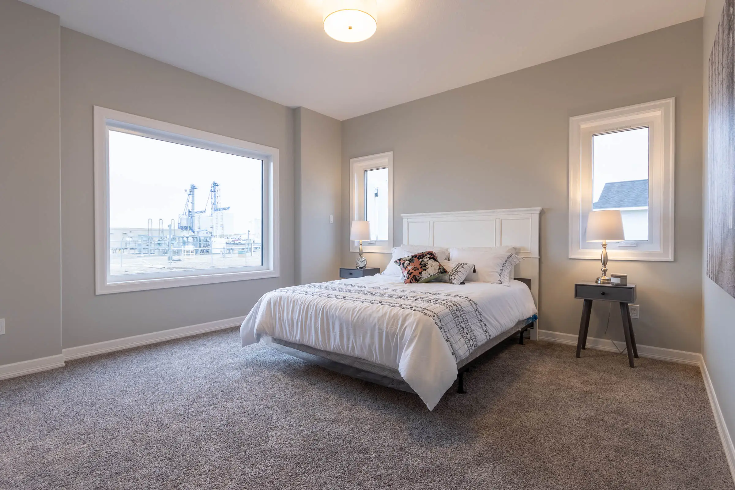 Spacious bedroom with gray walls, carpet, a white bed, and two bedside tables with lamps. Large windows reveal an industrial view outside.