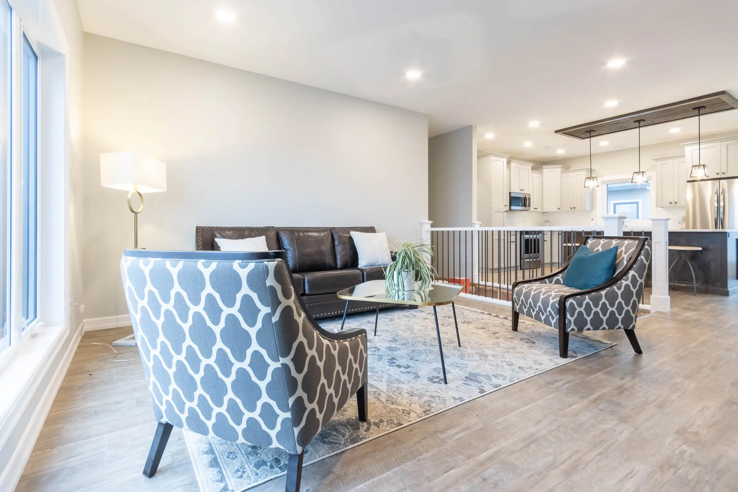 Modern living room with a dark sofa, patterned armchairs, glass coffee table, soft lighting, and open kitchen. The space is calm and inviting.