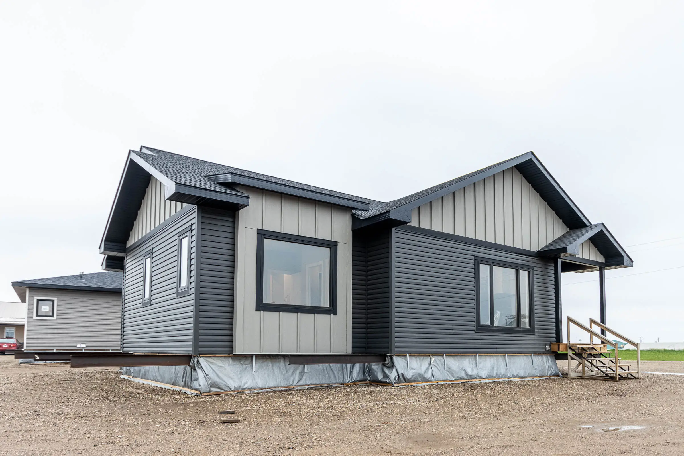 Modern modular home with gray siding and a gabled roof on a gravel lot. Large windows allow natural light. Overcast sky adds a muted tone.