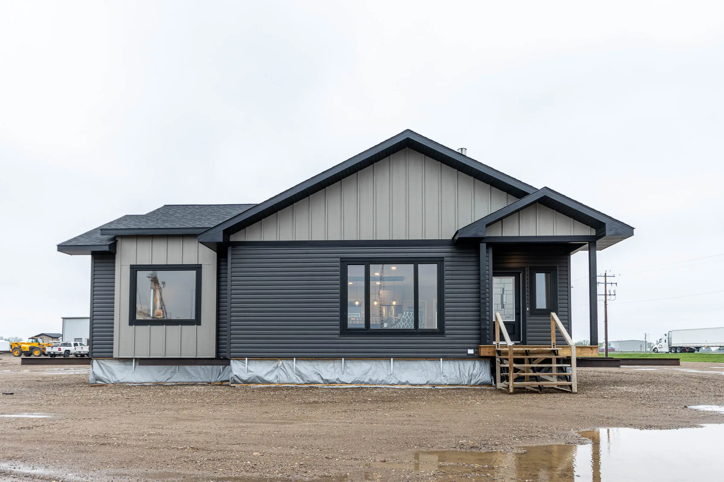 A single-story modular home with dark gray siding and steep gabled roof sits on a muddy lot. Wooden steps lead to the entrance, and large windows reveal a warmly lit interior.