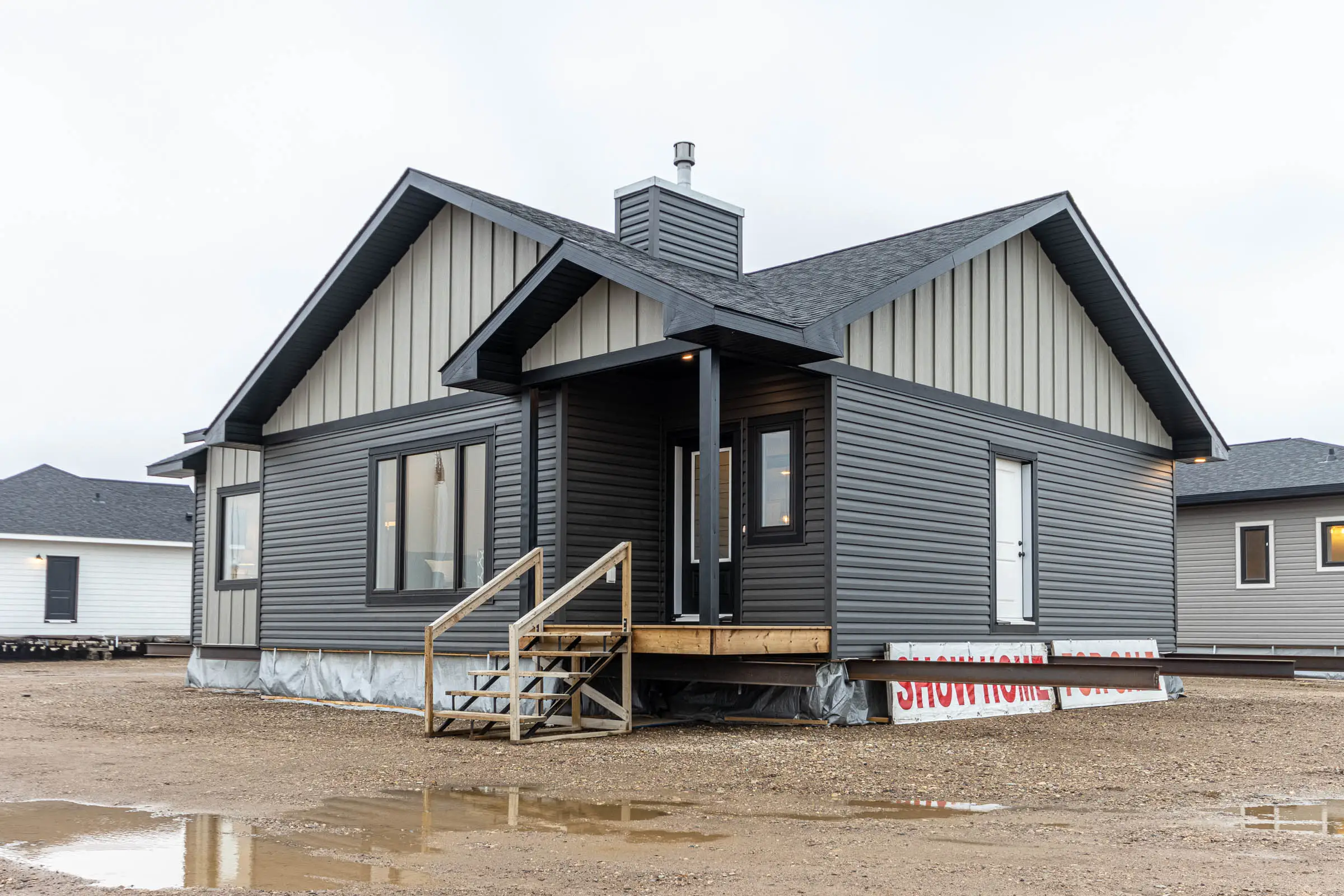 Single-story, gray modular home with dark siding and a small porch, set on a gravel lot. Overcast sky and puddles create a calm, muted atmosphere.