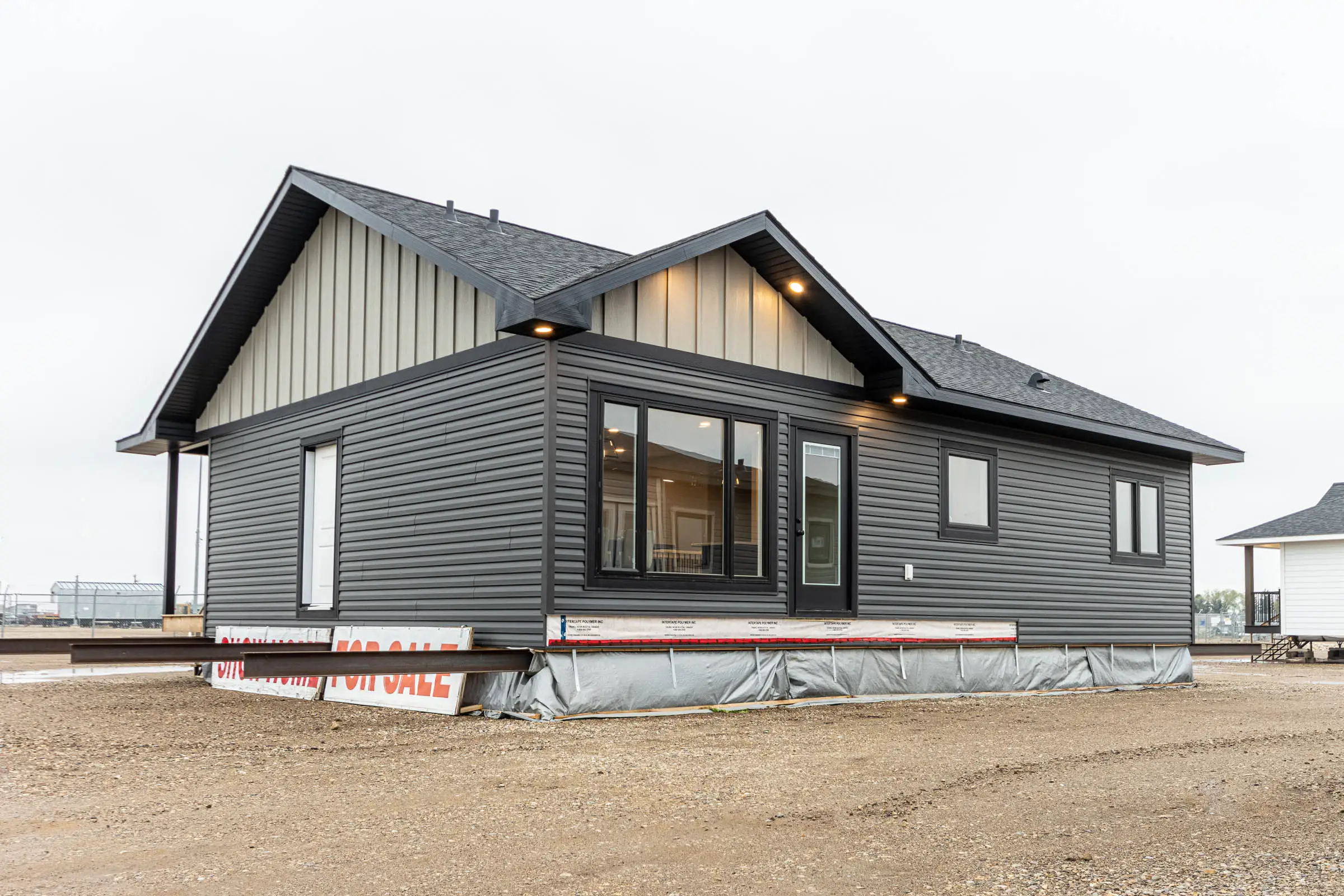 Modern, single-story prefabricated house with dark gray siding and a gabled roof. Situated on a dirt lot, a "For Sale" sign is visible at the base.