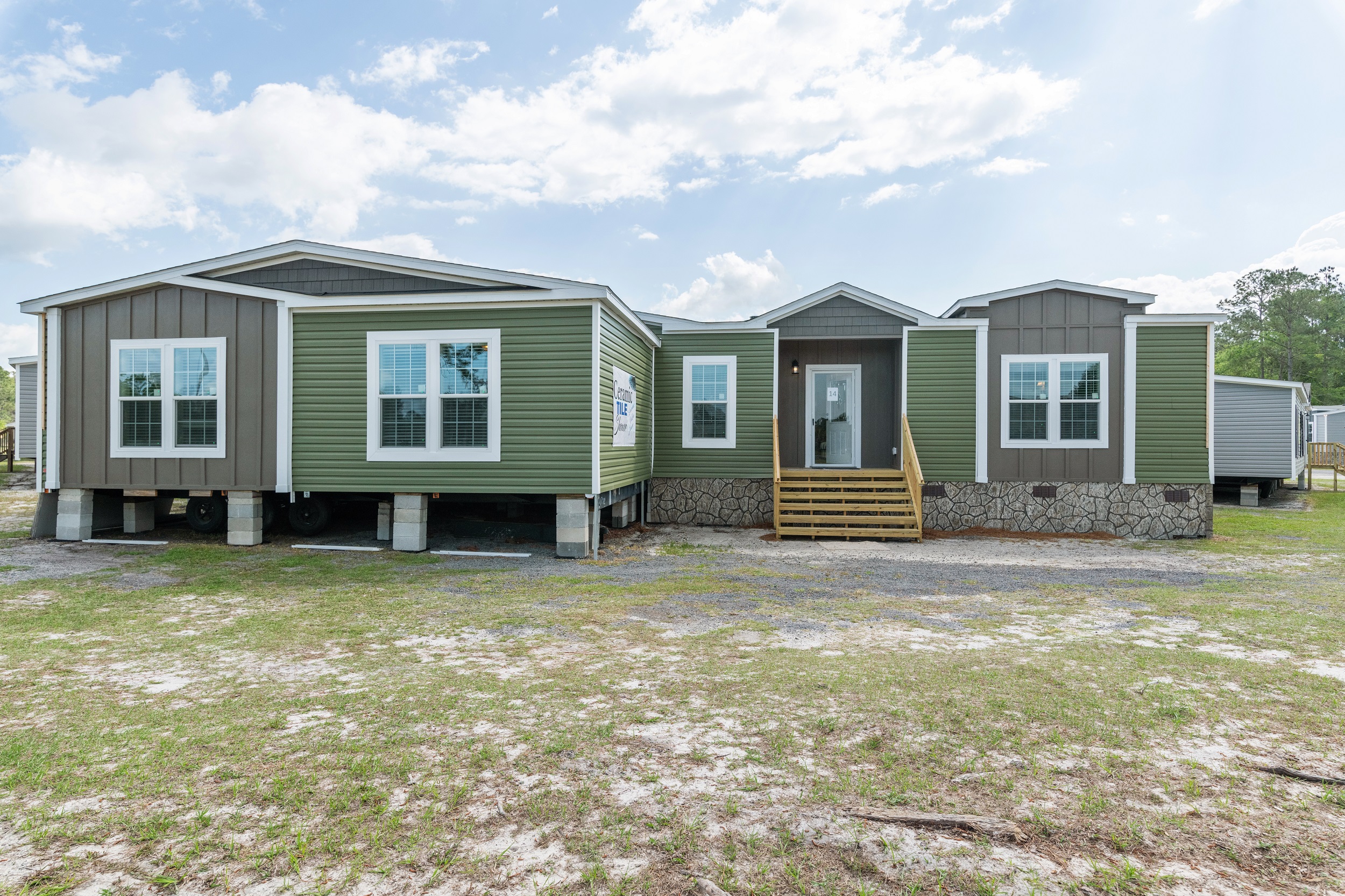 A modern, single-story modular home with green siding and white trim. It has several large windows, a central wooden staircase, and sits on a grassy lot under a partly cloudy sky.