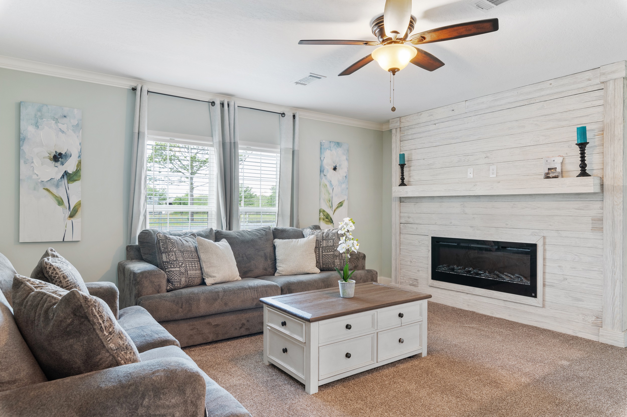 Stylish living room with gray sofas and soft cushions, centered around a wooden coffee table. A modern fireplace and floral artwork add coziness.