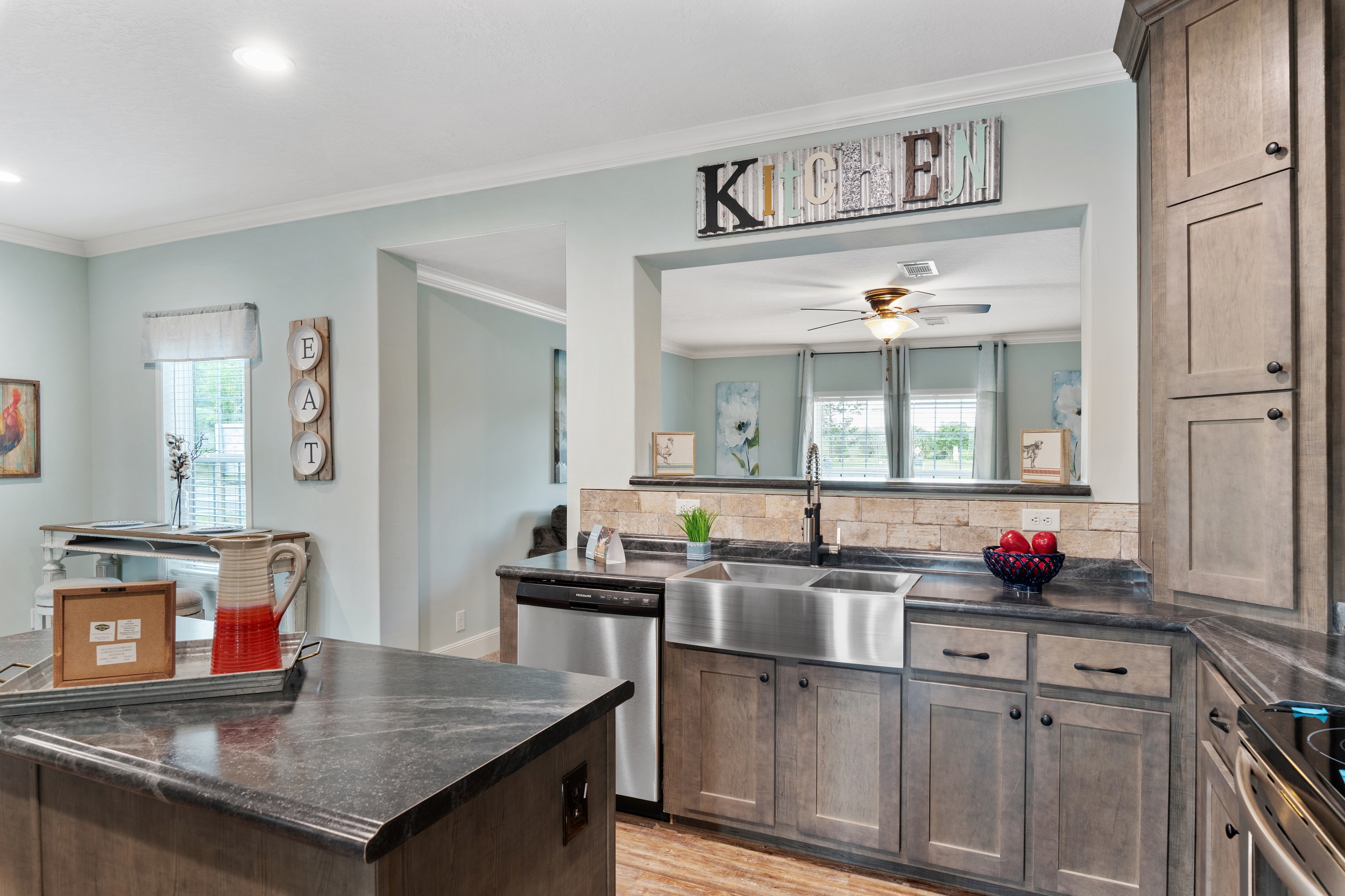 Modern kitchen with gray cabinets, stainless steel appliances, and a large farmhouse sink. A decorative "Kitchen" sign hangs above, conveying a welcoming ambiance.