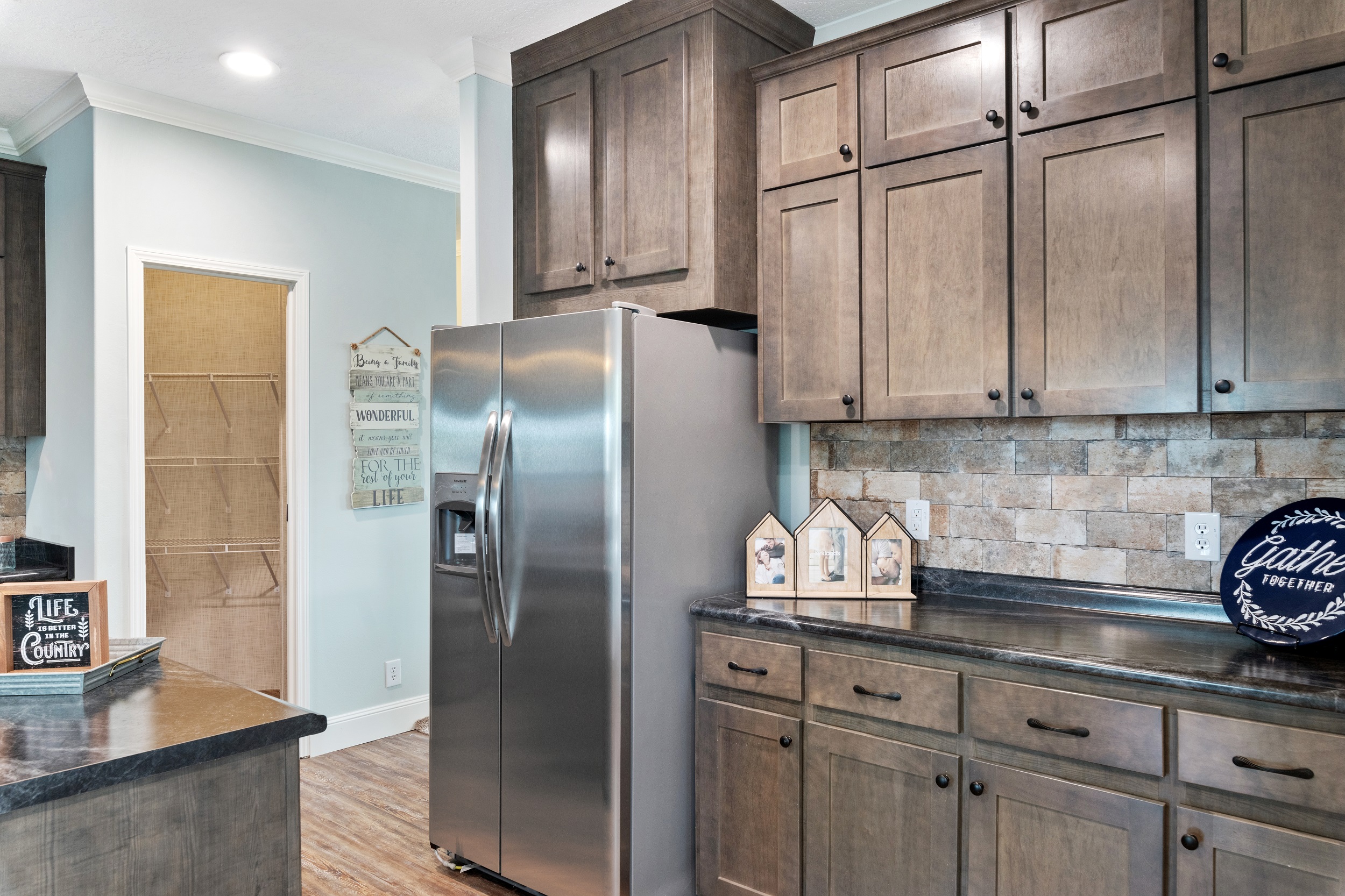 Spacious kitchen with dark wood cabinets, stainless steel fridge, tile backsplash, and black countertops. Cozy decor includes a "Gather" plate and small wooden houses.