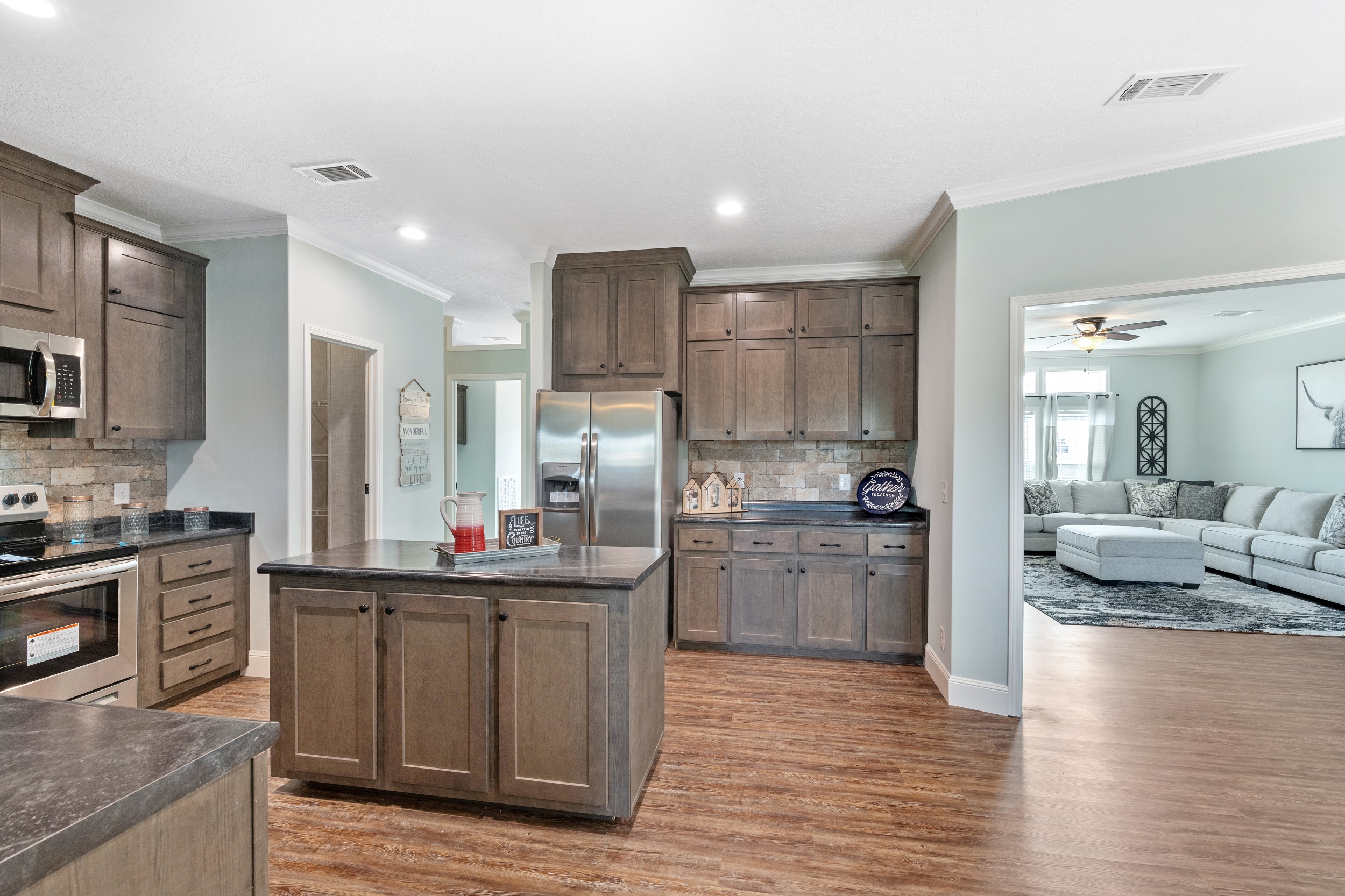 Modern kitchen with dark wood cabinets, central island, and stainless steel appliances on wooden flooring. Open view to a cozy, bright living room.