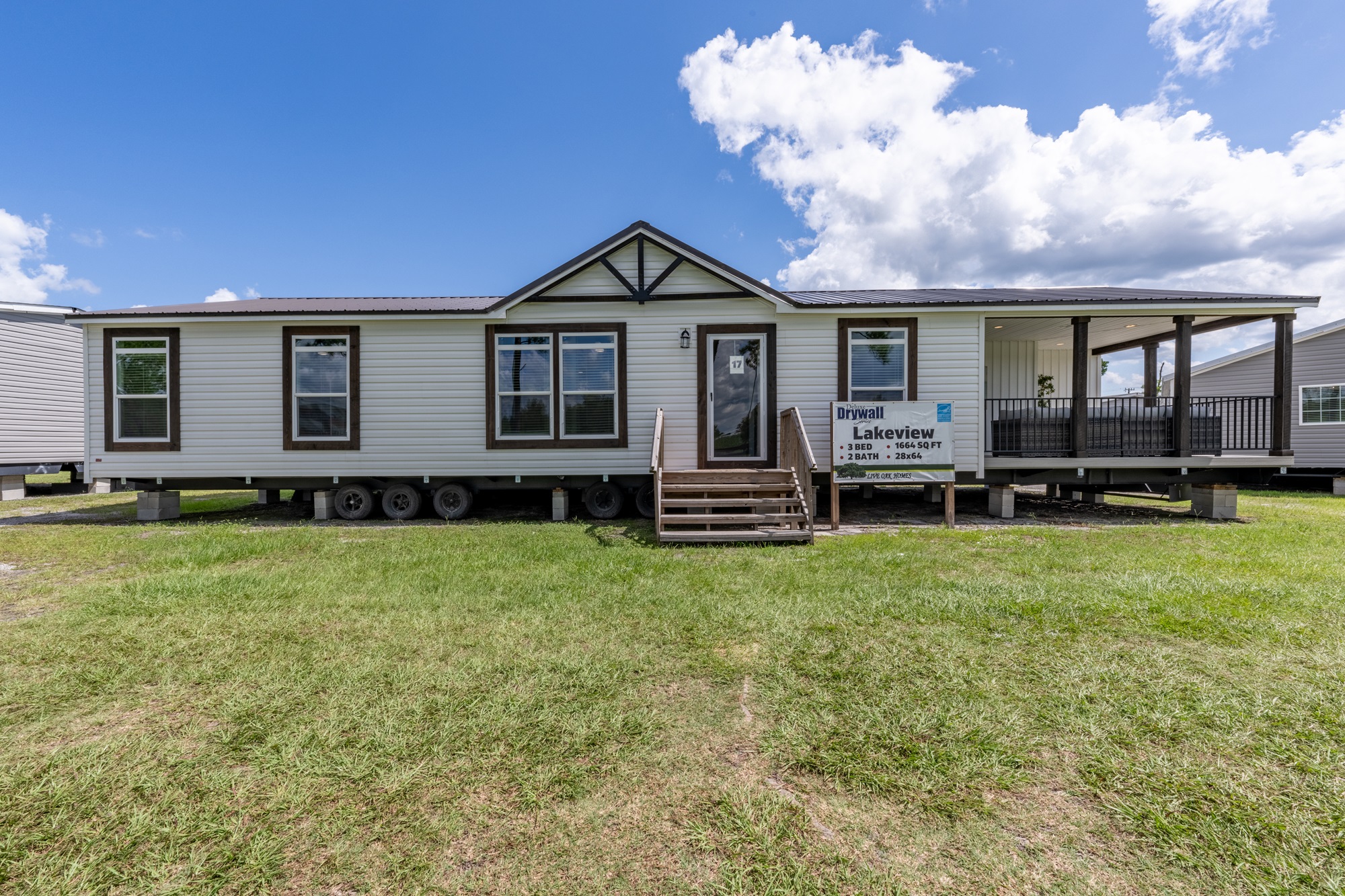 A modern mobile home with a gray exterior, brown trim, and a covered porch. It sits on a grassy lot under a blue sky with scattered clouds.
