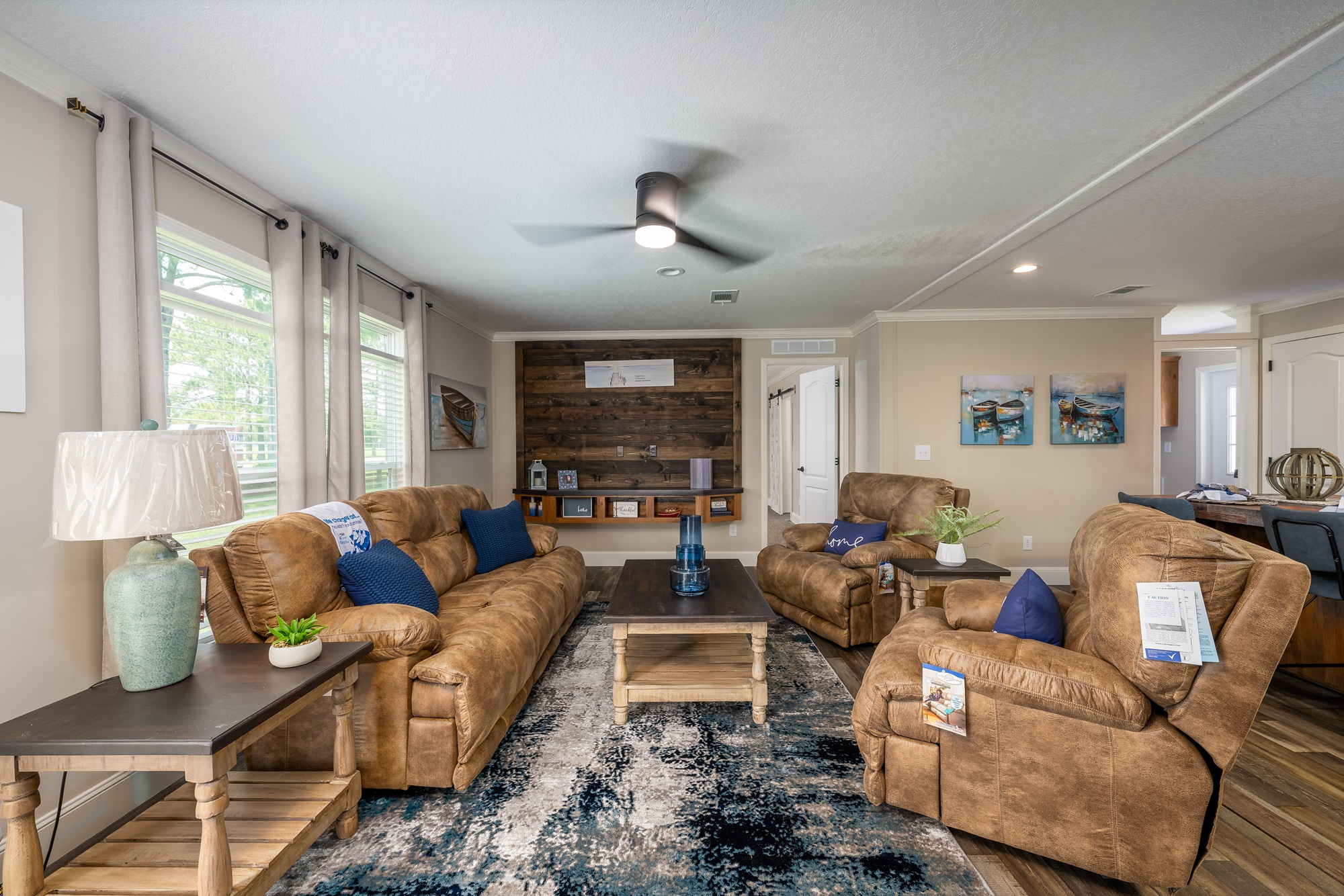 Cozy living room with brown recliners and sofas, accented by blue cushions. A wooden coffee table sits on a blue patterned rug. A wooden feature wall and decor add warmth, while large windows provide natural light.