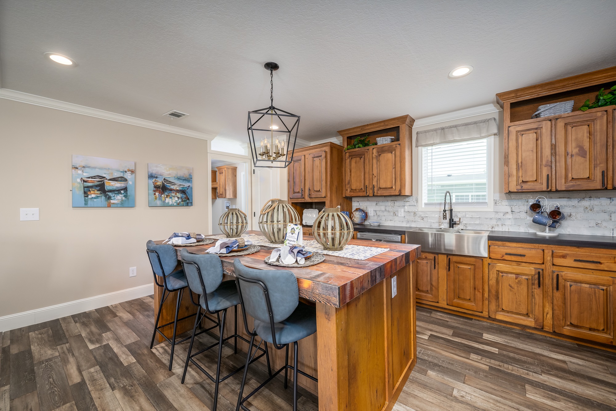 Warm kitchen with wooden cabinets, a central island with four gray chairs, and a chandelier. Bright, welcoming ambiance with decorative accents.
