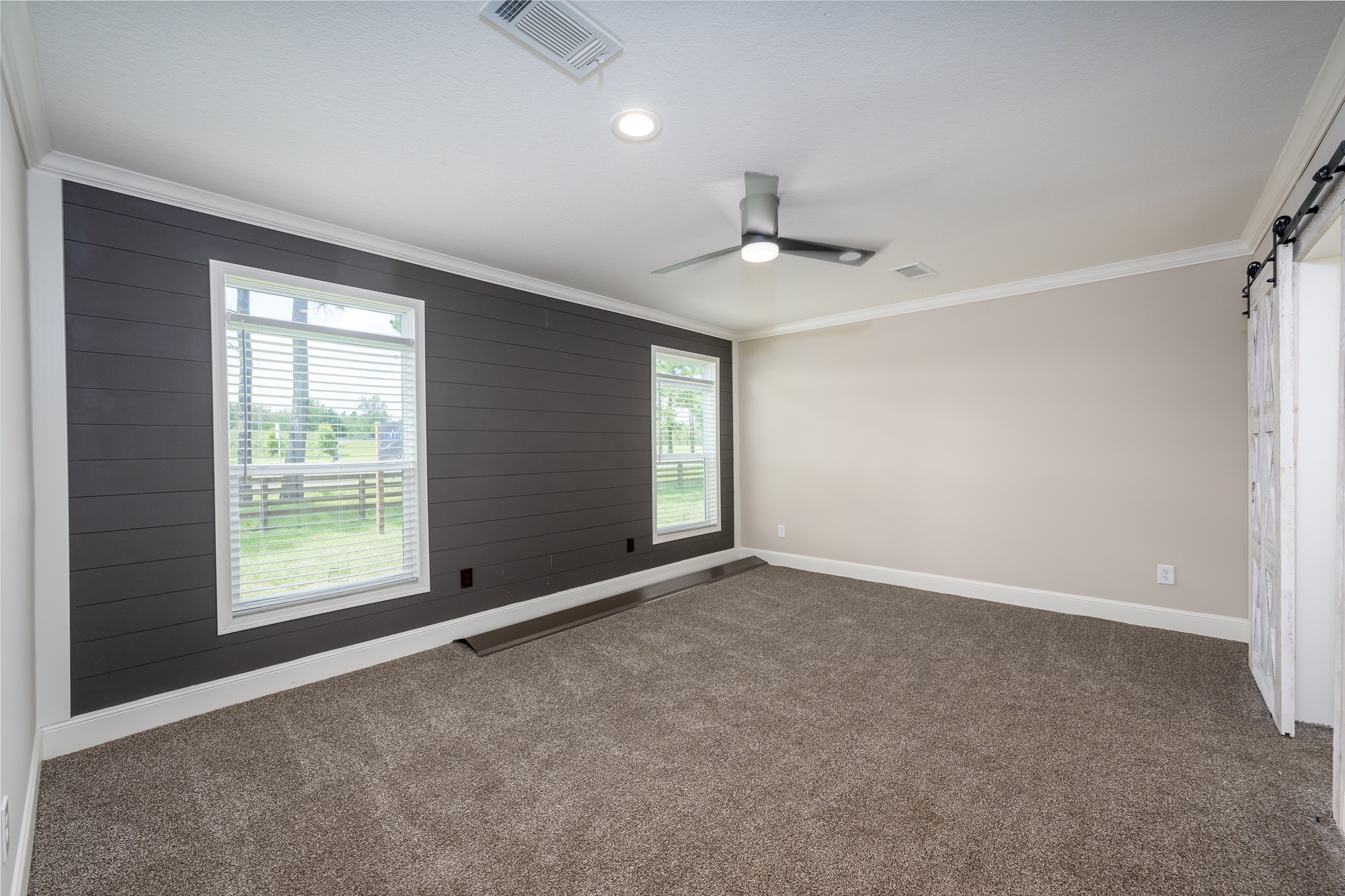 Spacious room with beige carpet, gray shiplap accent wall, and two large windows. Ceiling fan and bright overhead lighting add a modern touch.