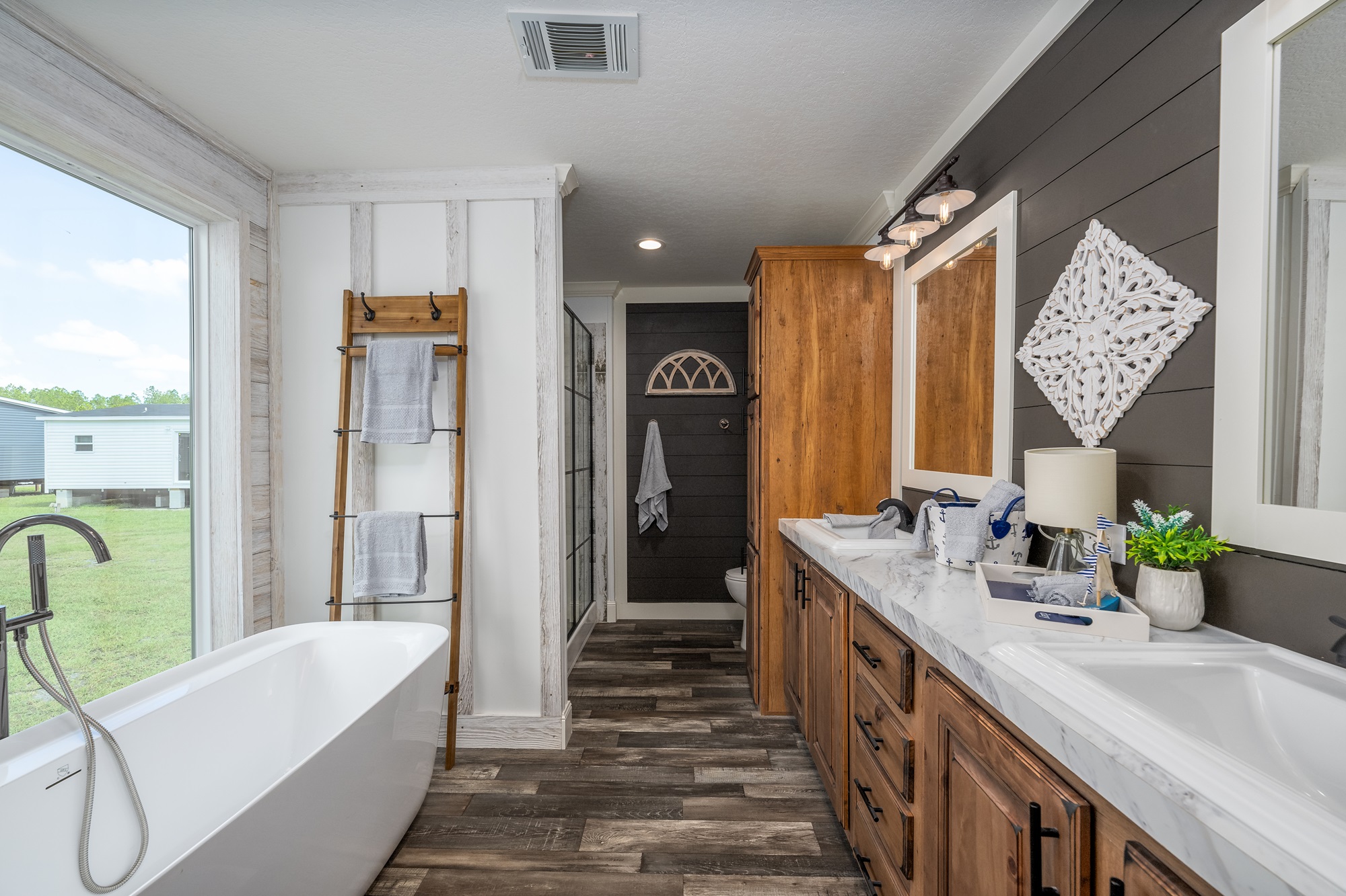 Modern bathroom with a freestanding tub near a large window, wood ladder towel rack, double vanity with marble top, and dark accent wall. Cozy and inviting.