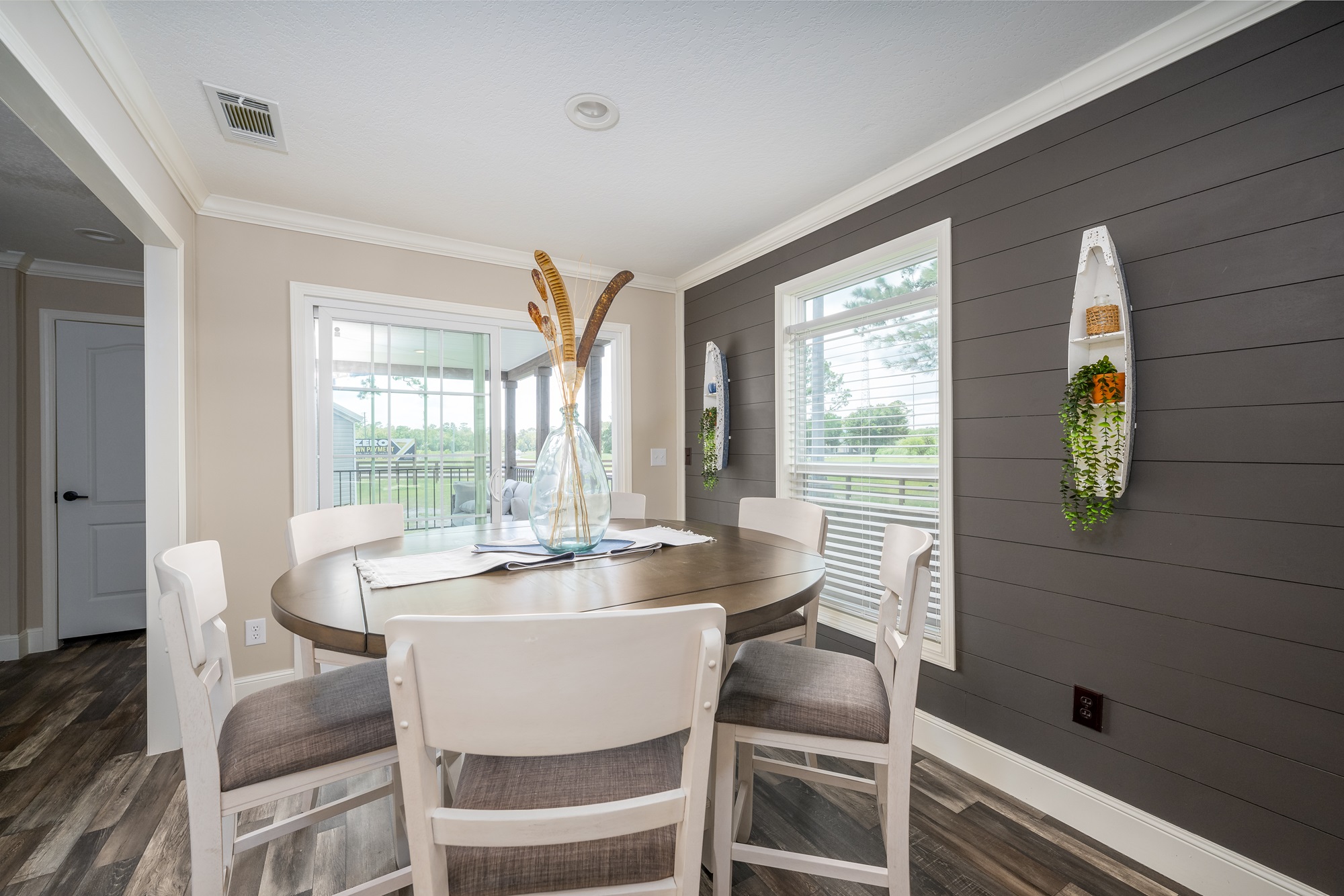 A cozy dining area with a round wooden table and six chairs against a gray shiplap wall. Large windows offer natural light. A vase with decorative branches sits on the table, and small planters hang beside the windows, adding a touch of greenery. The tone is inviting and modern.