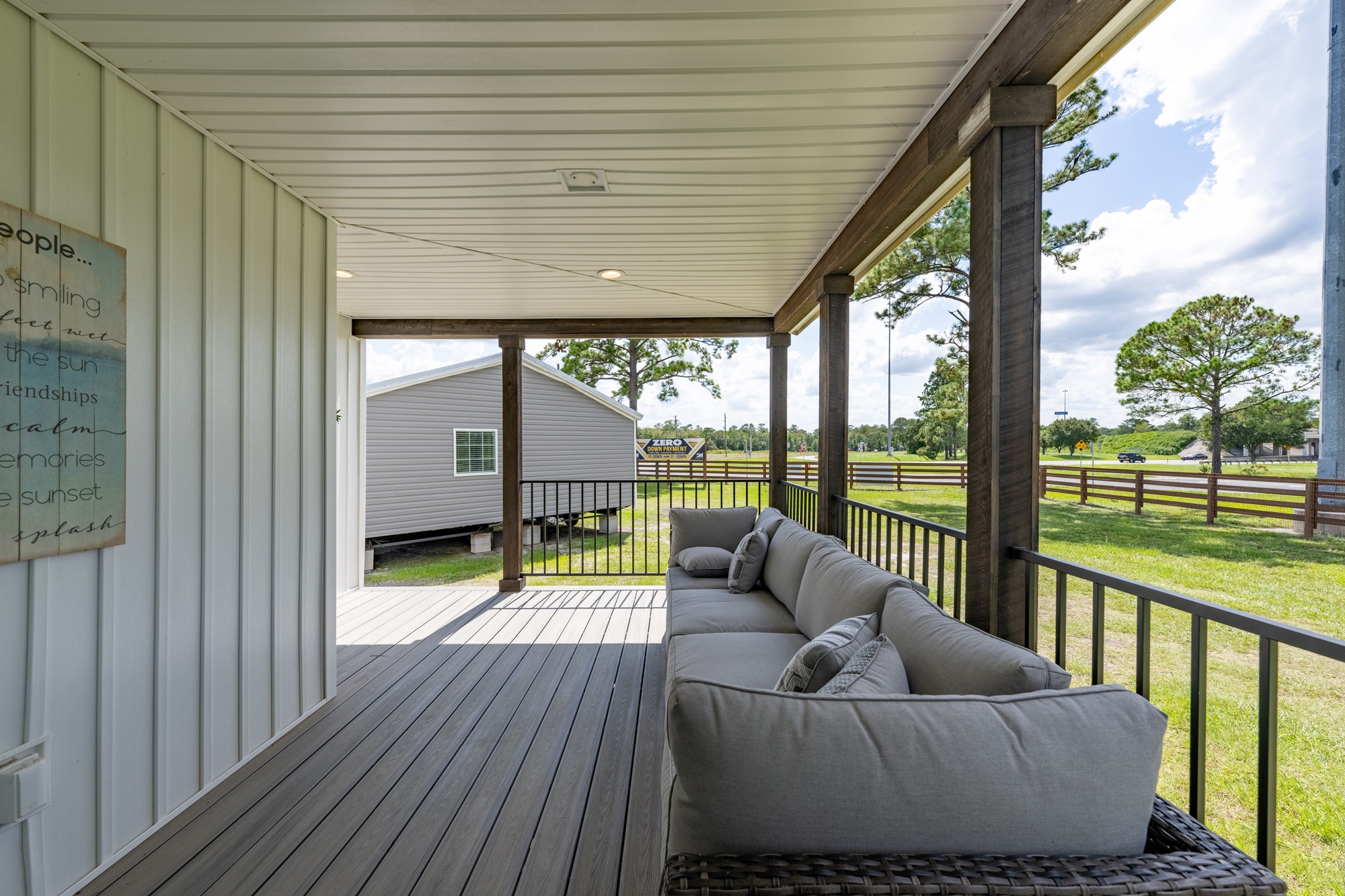 A cozy porch with a wicker sofa adorned with gray cushions. It overlooks a grassy yard with trees and a view of a nearby building and fence.