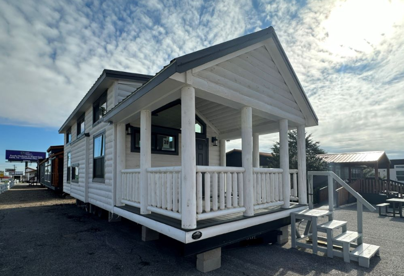 Alt text: "A small log-style cabin with a white wooden front porch and steps sits under a partly cloudy sky. The scene conveys a peaceful, rustic charm."