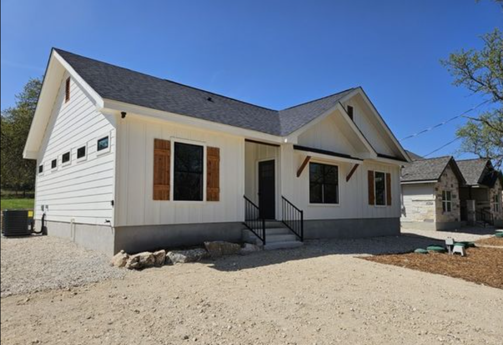 Single-story white house with dark roof, wooden shutters, and front porch under clear blue sky. Gravel yard and a neighboring house visible on the right.