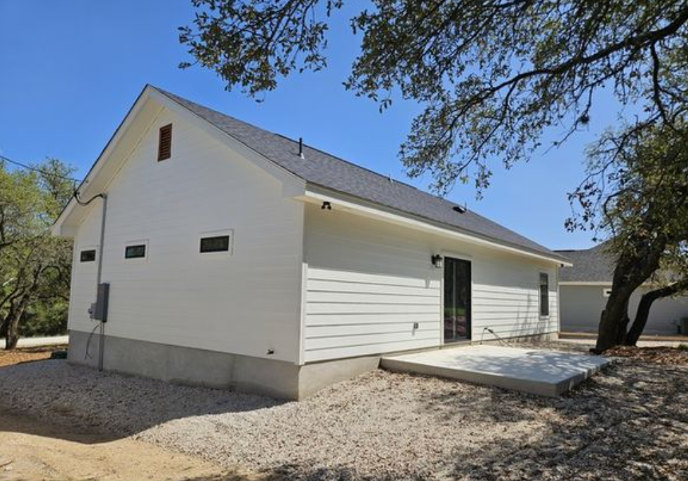 A small white house with a dark gray roof sits in a yard with gravel and trees. A small patio is visible, and the scene conveys simplicity and tranquility.