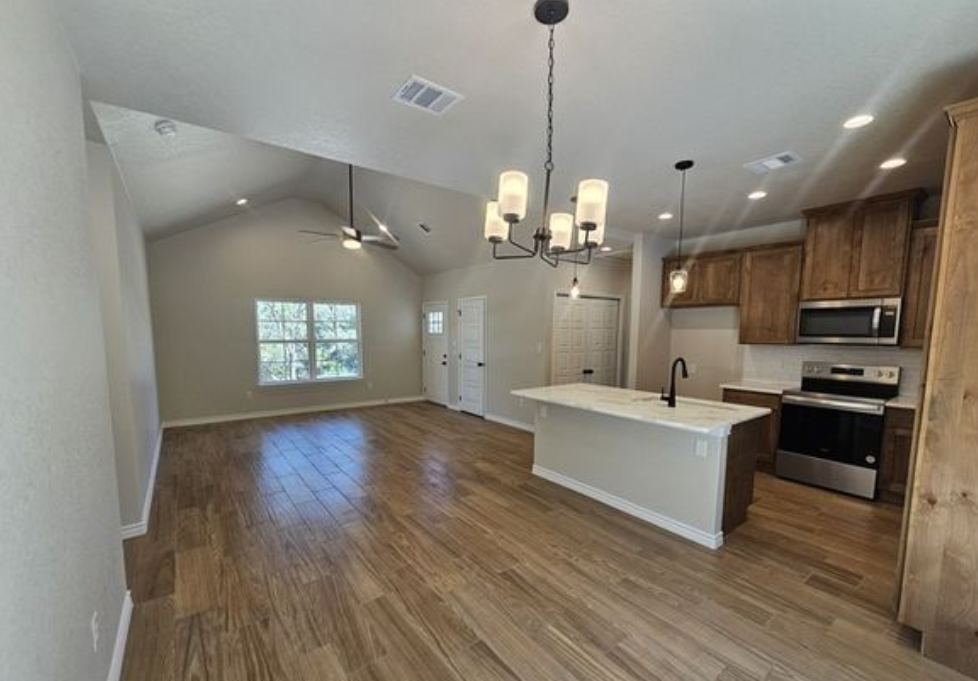 Spacious open-concept kitchen and living area with vaulted ceiling and wood flooring. Features include an island, stainless steel appliances, and wooden cabinets. Warm, inviting lighting from hanging fixtures enhances the cozy ambiance.