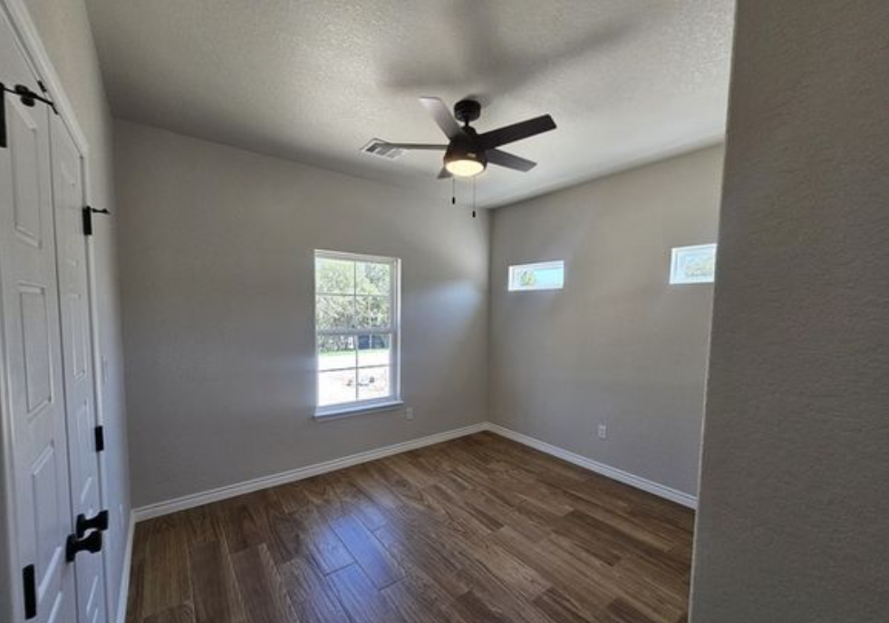 A small, empty room with wooden floors, light gray walls, and three windows. A ceiling fan is centered above, evoking a calm, minimalist vibe.