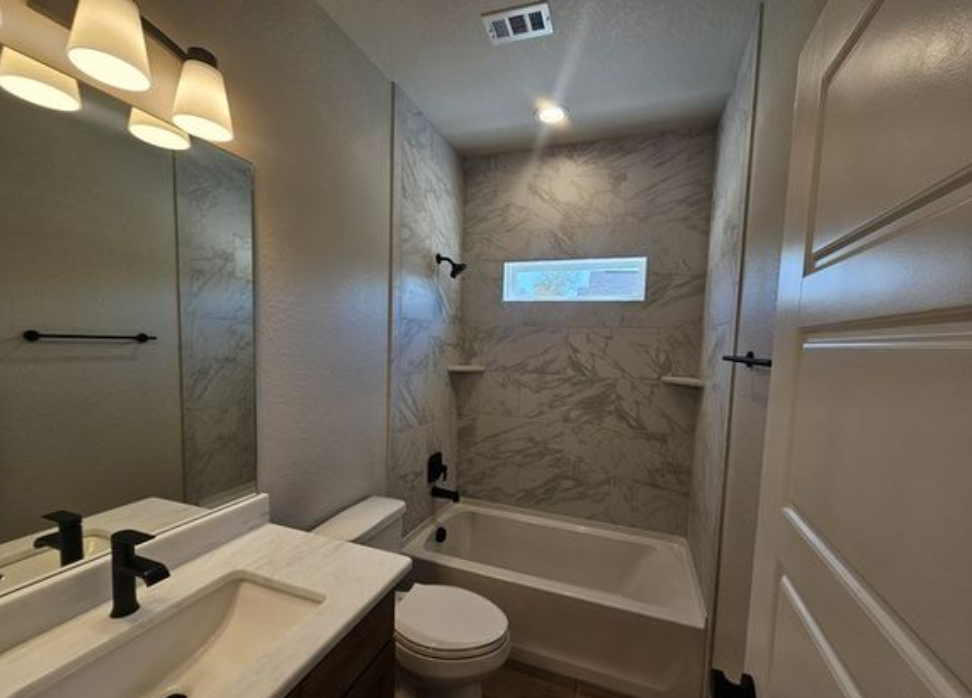 Modern bathroom with marble-patterned shower walls, a white bathtub, black fixtures, a rectangular mirror, and three pendant lights. Soft, neutral tones convey elegance.