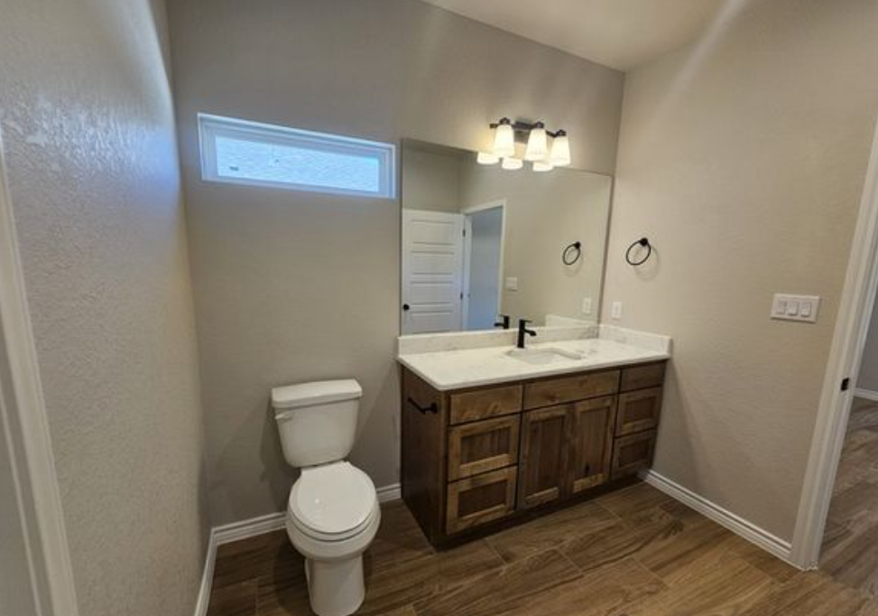 Simple bathroom with wooden vanity, white countertop, and white toilet. Modern light fixture and rectangular mirror add elegance. Neutral tones.