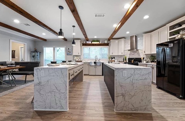 Spacious kitchen with marble islands, wooden beams, and pendant lights. Stainless steel appliances and large window add a modern, airy feel.
